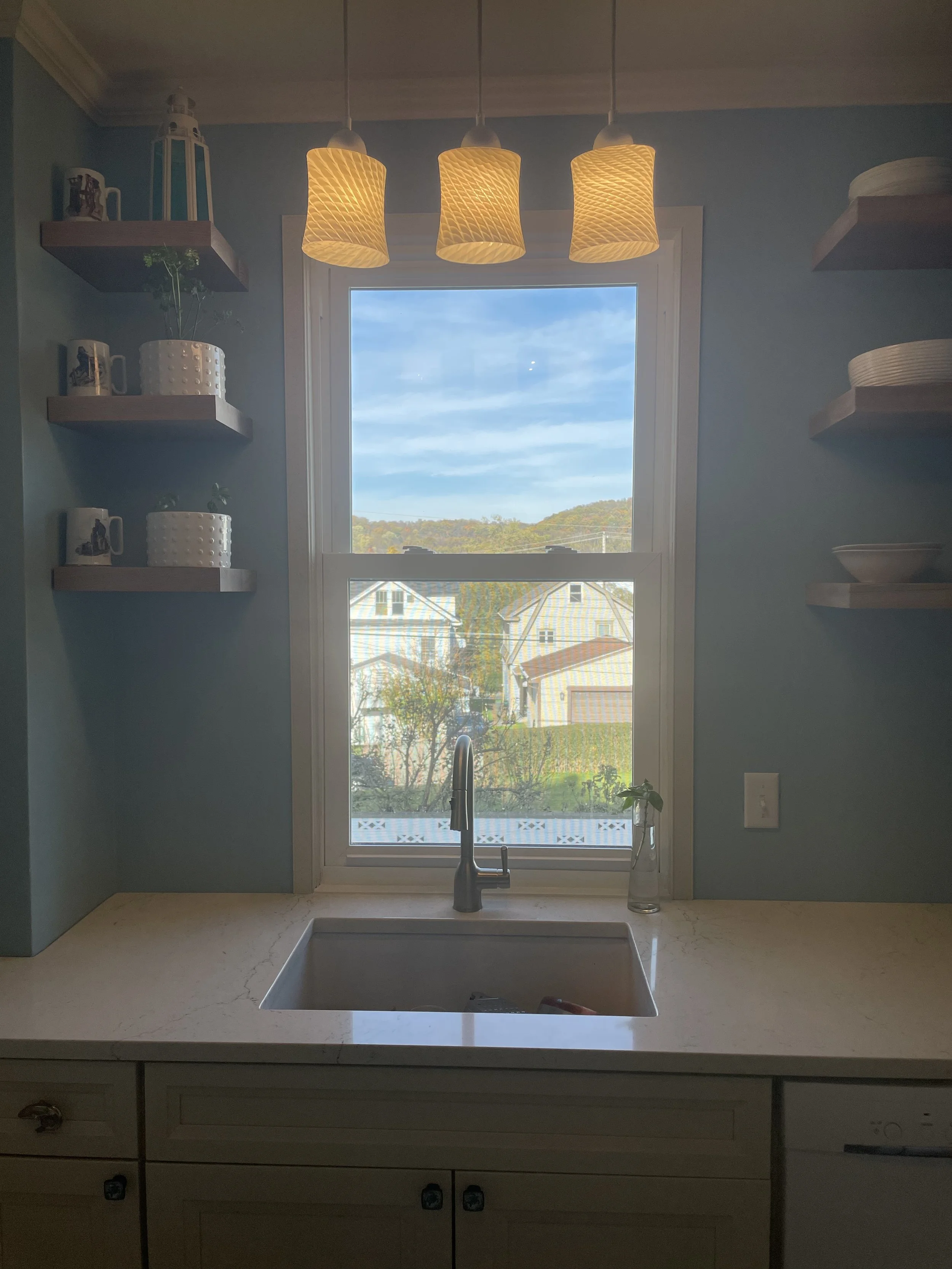 Kitchen sink beneath a window with a view of houses and trees outside. There are three pendant lights hanging above, and shelves with dishes and decorative items on each side.