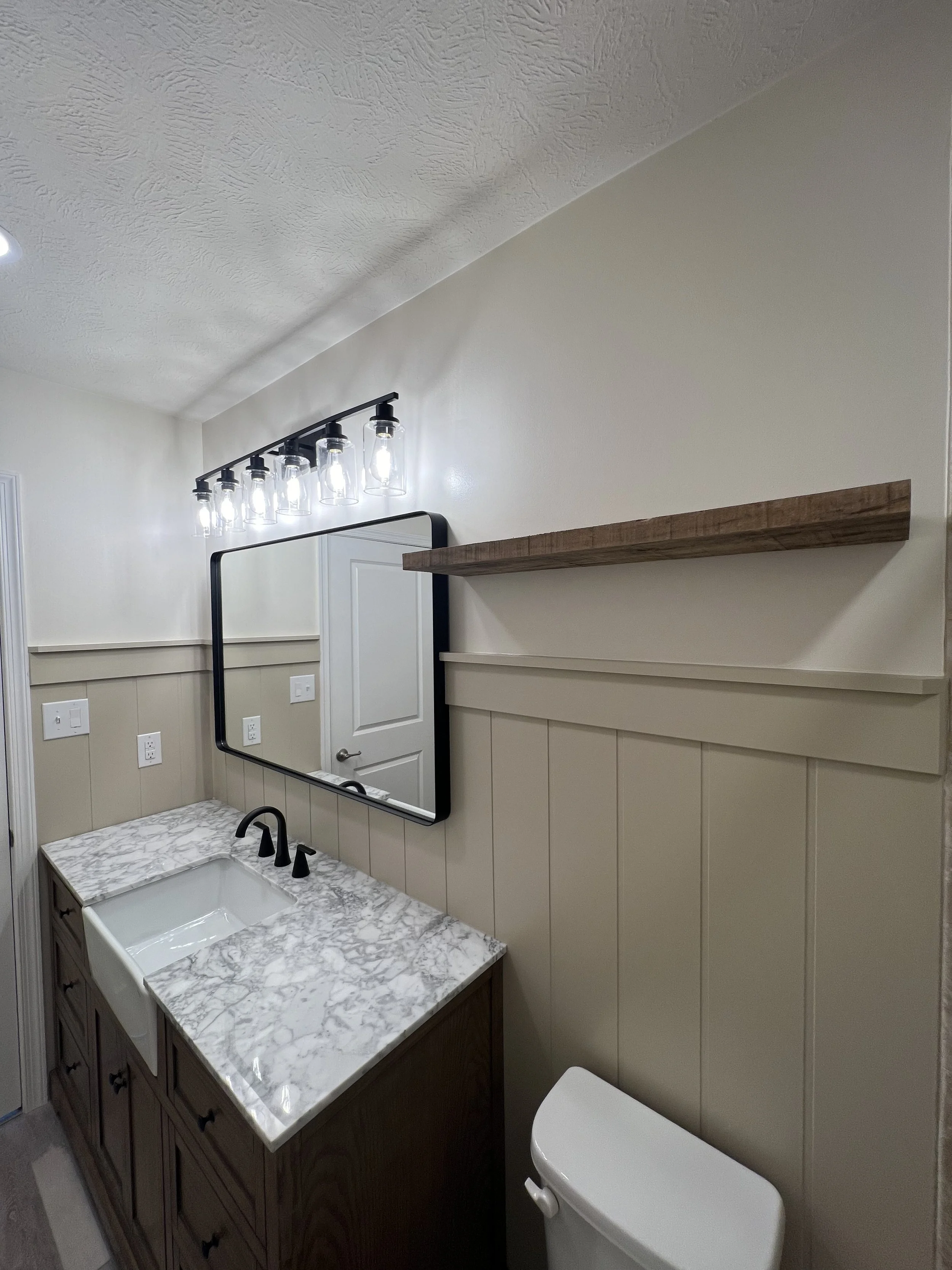 Bathroom with a marble countertop sink, a black faucet, a wall mirror, a wood shelf, beige beadboard wall paneling, and a white toilet.
