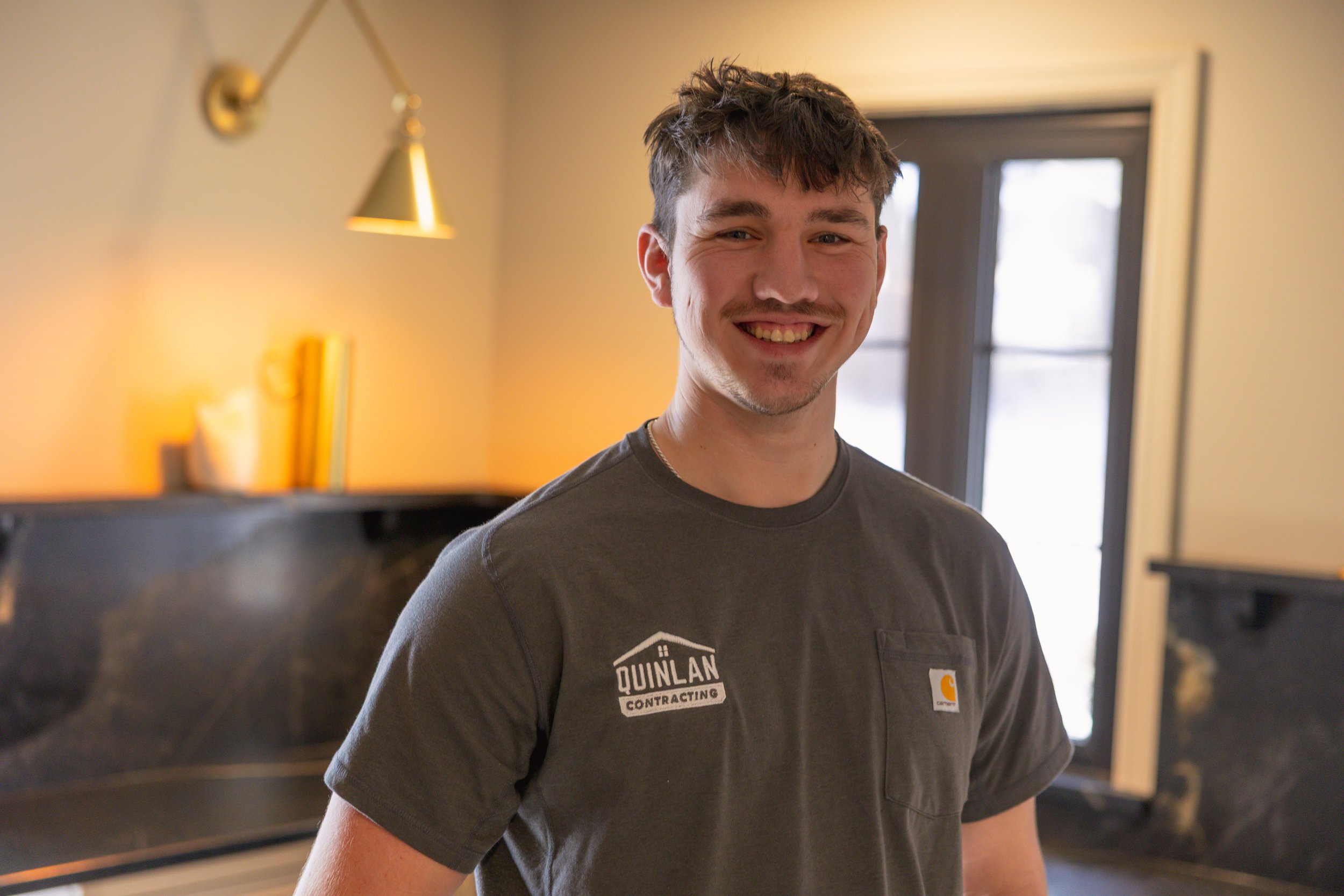 A young man with short brown hair and a beard smiling in a warmly lit room, wearing a dark gray Carhartt t-shirt with a logo, standing in front of a window with black framing.