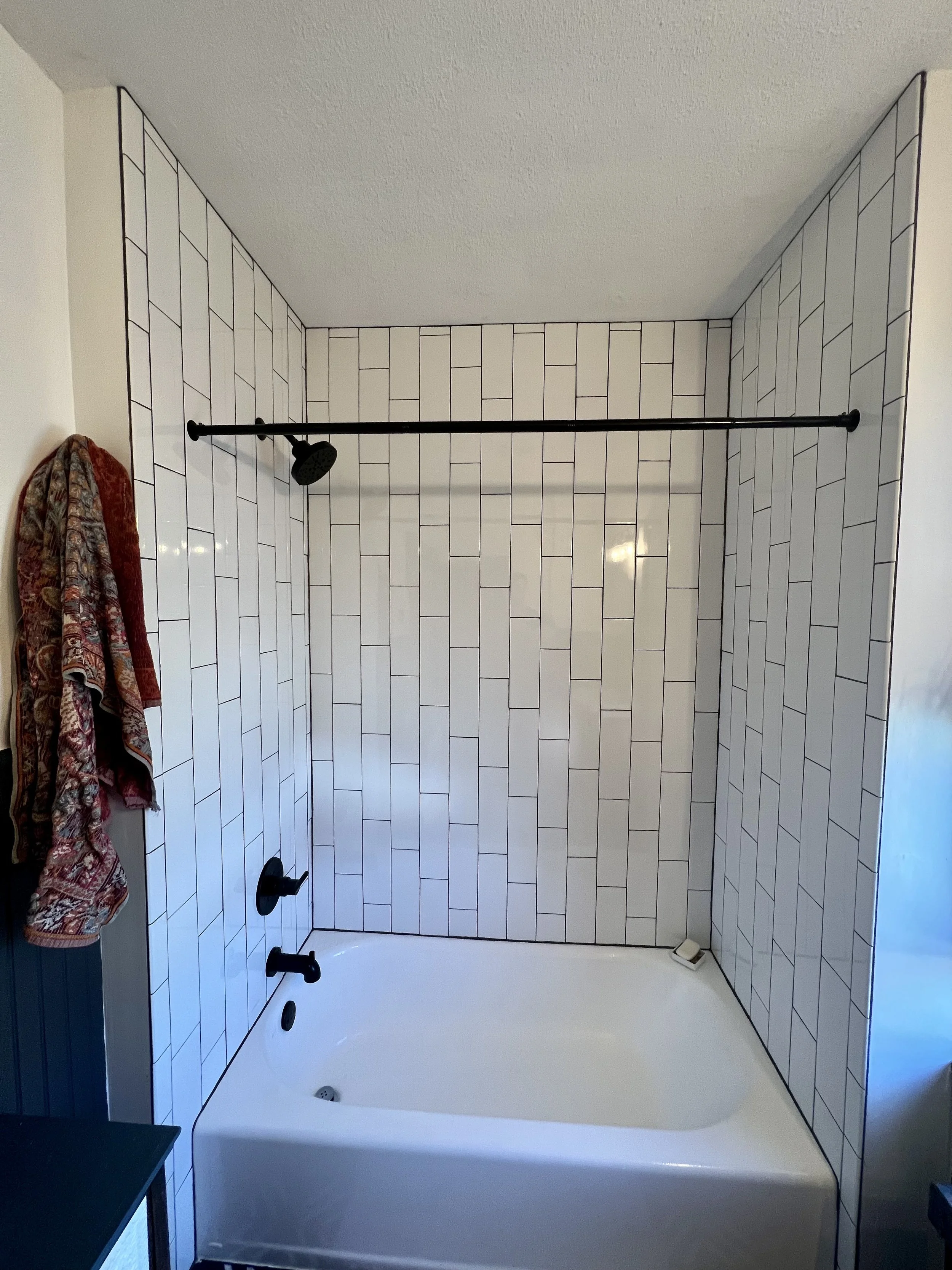 A bathroom with white tile walls, a bathtub, a black shower head, and a black faucet. A red and patterned towel hangs on the left wall.