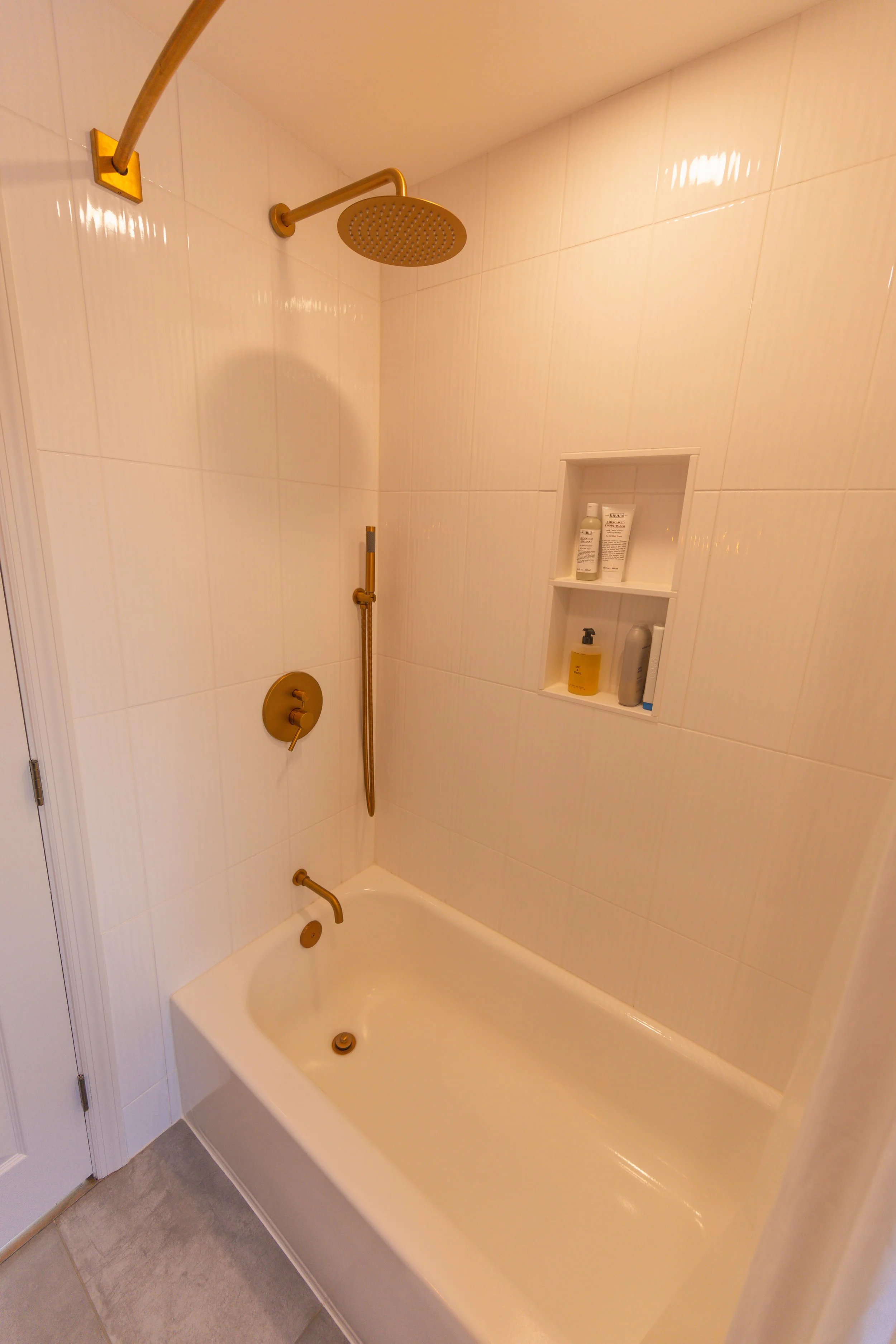 Bathroom shower with white tiled walls, gold fixture, mid-sized showerhead, and built-in shelf with toiletries.