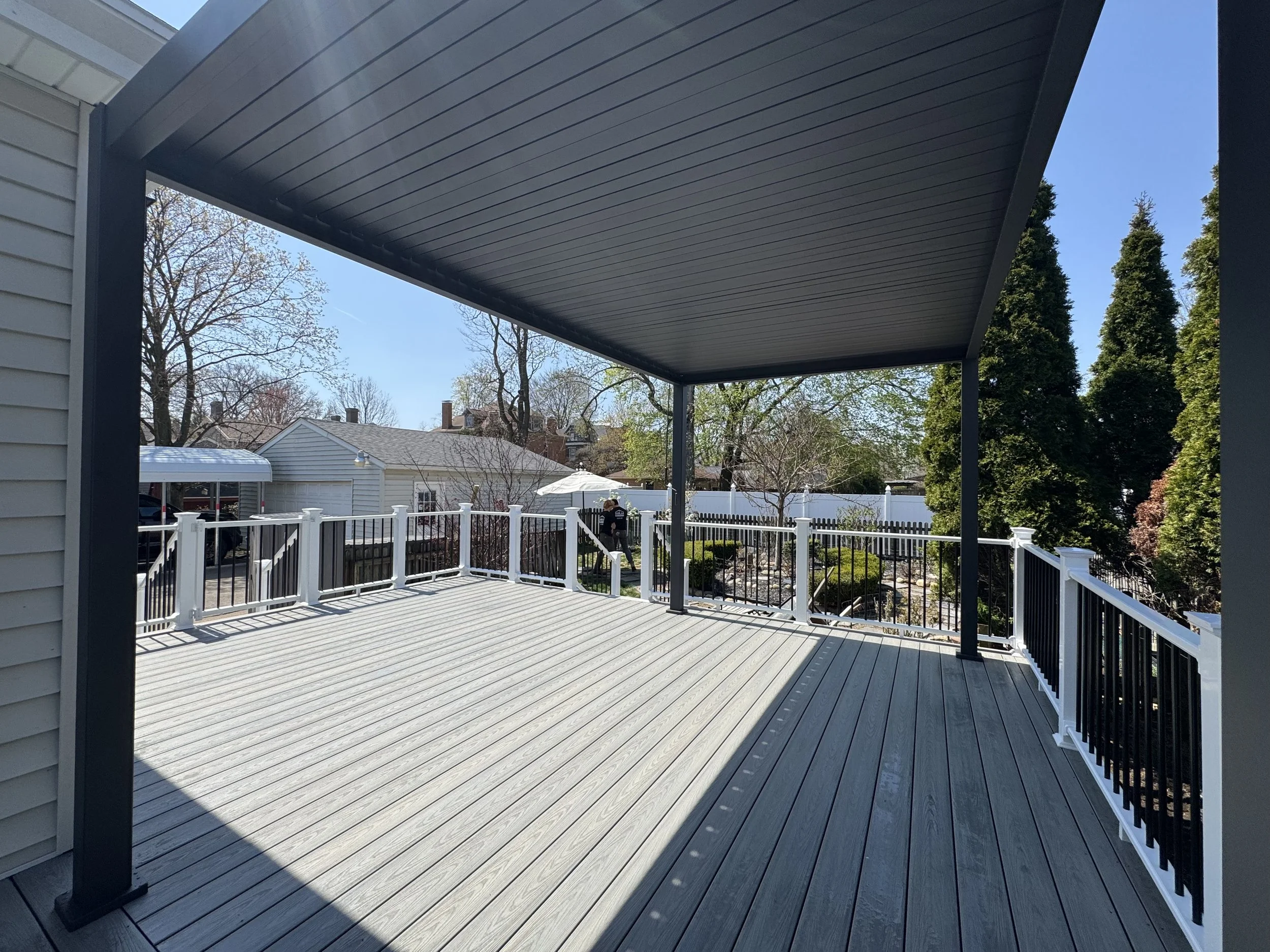 View of a spacious outdoor deck with a black and white railing, covered by a dark-colored roof, overlooking neighboring houses and tall trees on a sunny day.