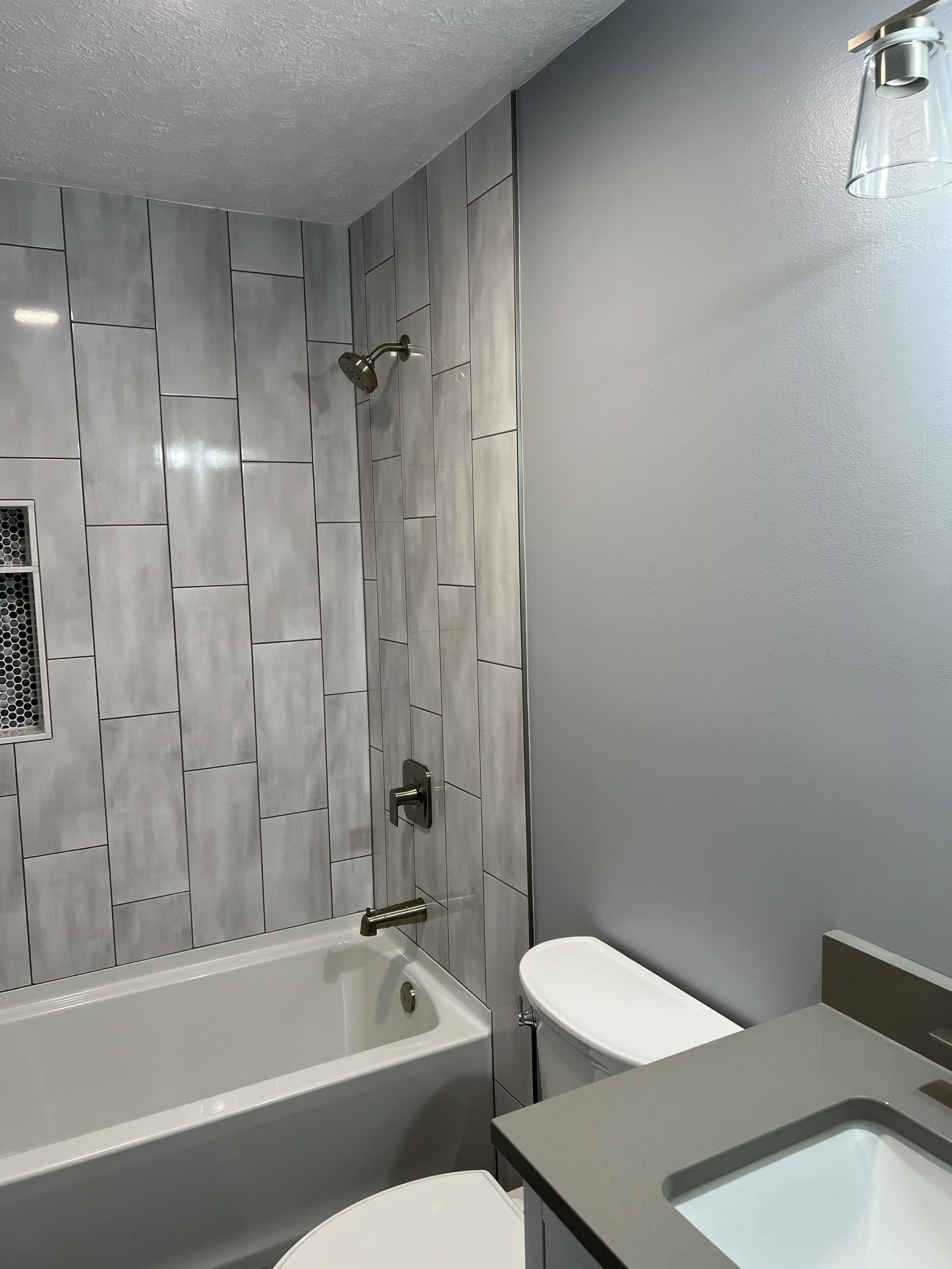 Modern bathroom featuring a bathtub with a showerhead, gray tiled wall, toilet, gray countertop, and a small sink.