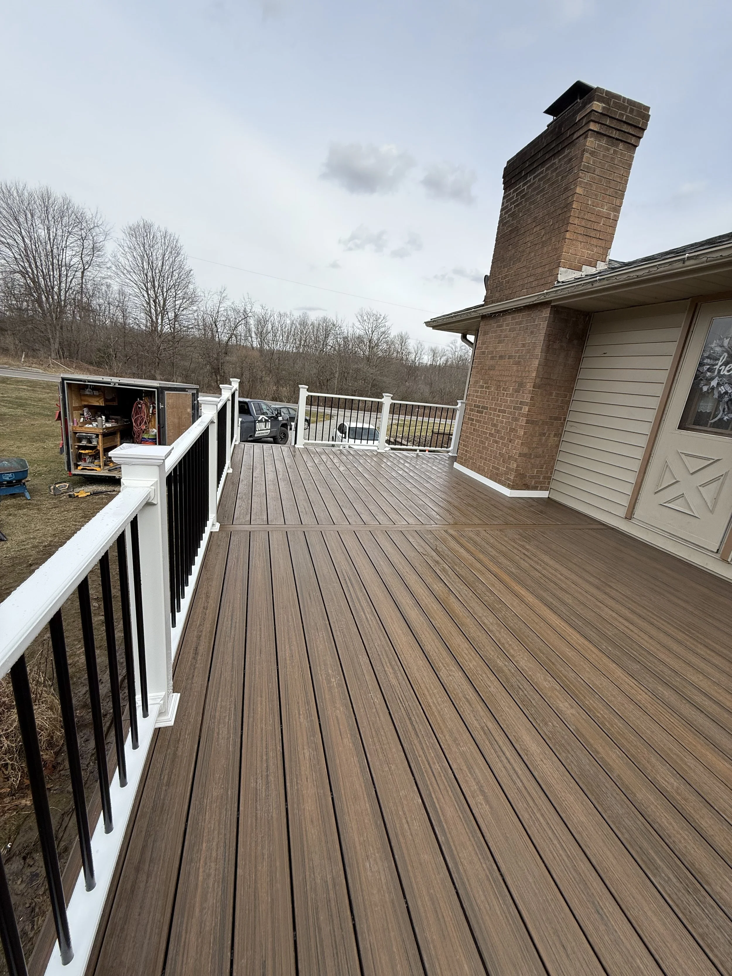 A newly installed wooden deck with railings, attached to a house with a brick chimney and a door decorated for the holidays. The surrounding yard has leafless trees, vehicles, and a tool shed.