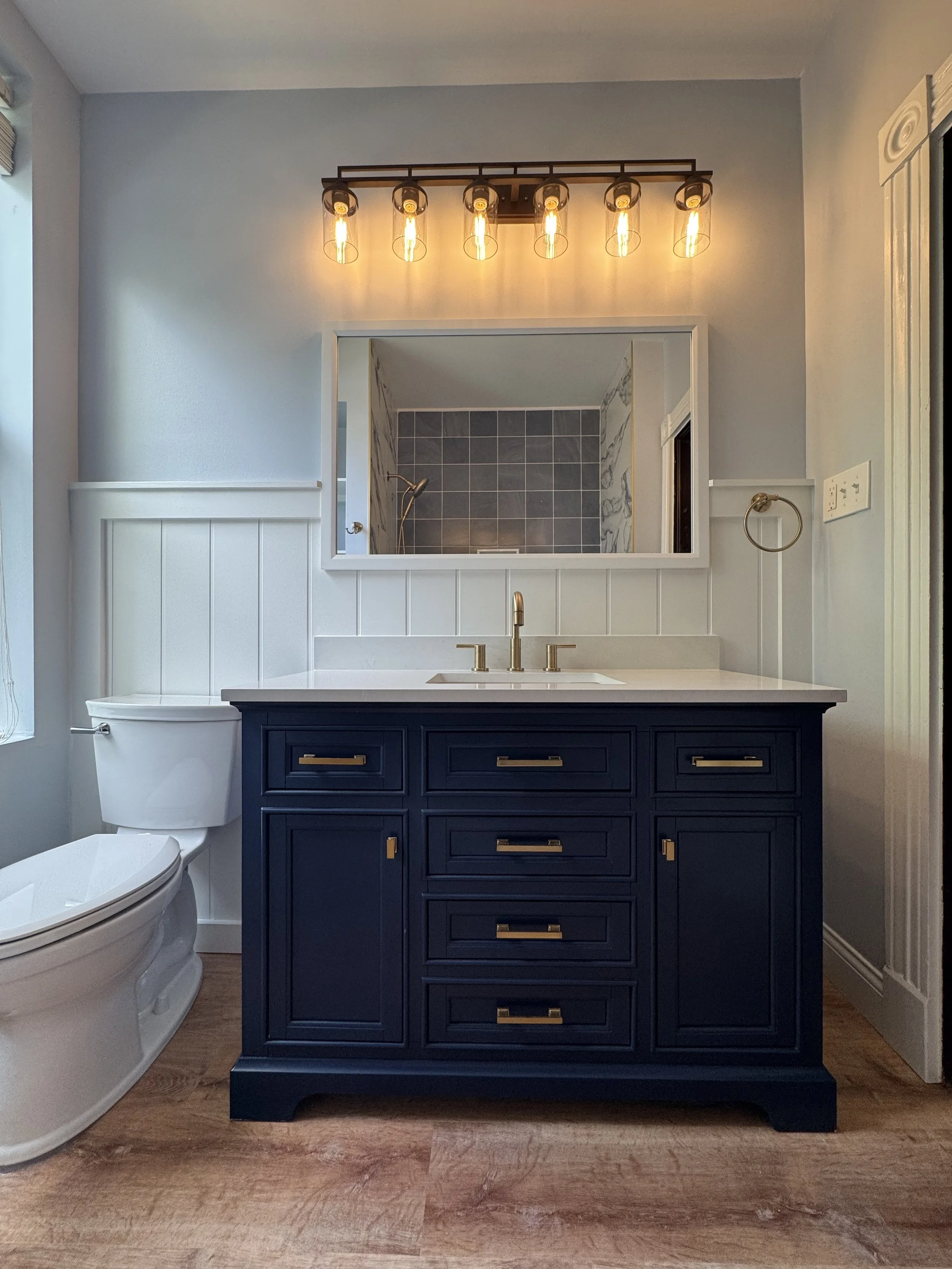 Bathroom with navy blue vanity, white countertop, and gold hardware. A mirror is above the vanity, reflecting a shower with gray tiles. To the left is a white toilet, and above it is wood paneling on the wall. Above the mirror is a vintage-style ligh