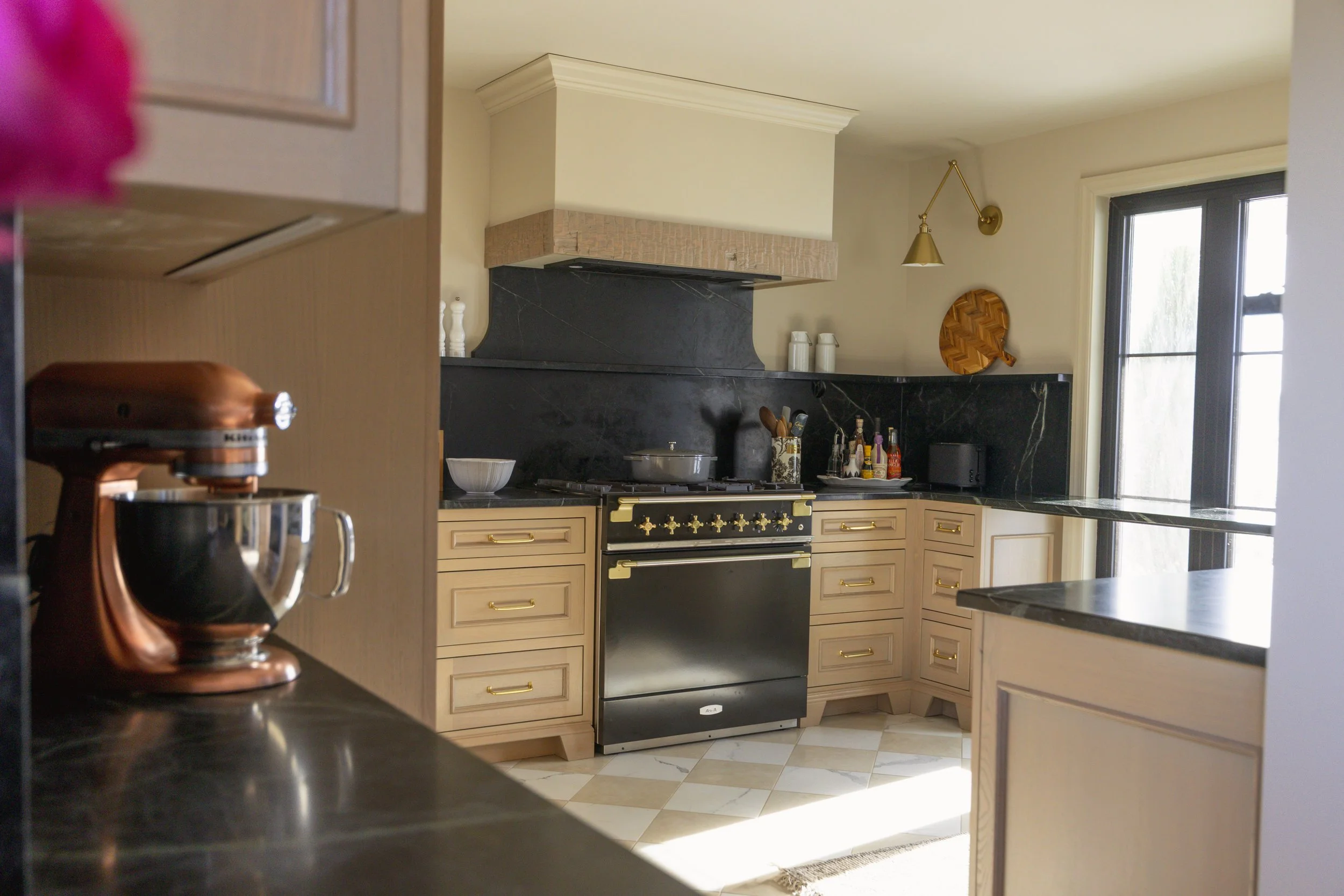 Kitchen with beige cabinets, black marble countertops, black stove, and black backsplash. There is a copper stand mixer on the counter to the left, and various kitchen items on the counters near the stove including a bowl, utensils, and bottles. A wi