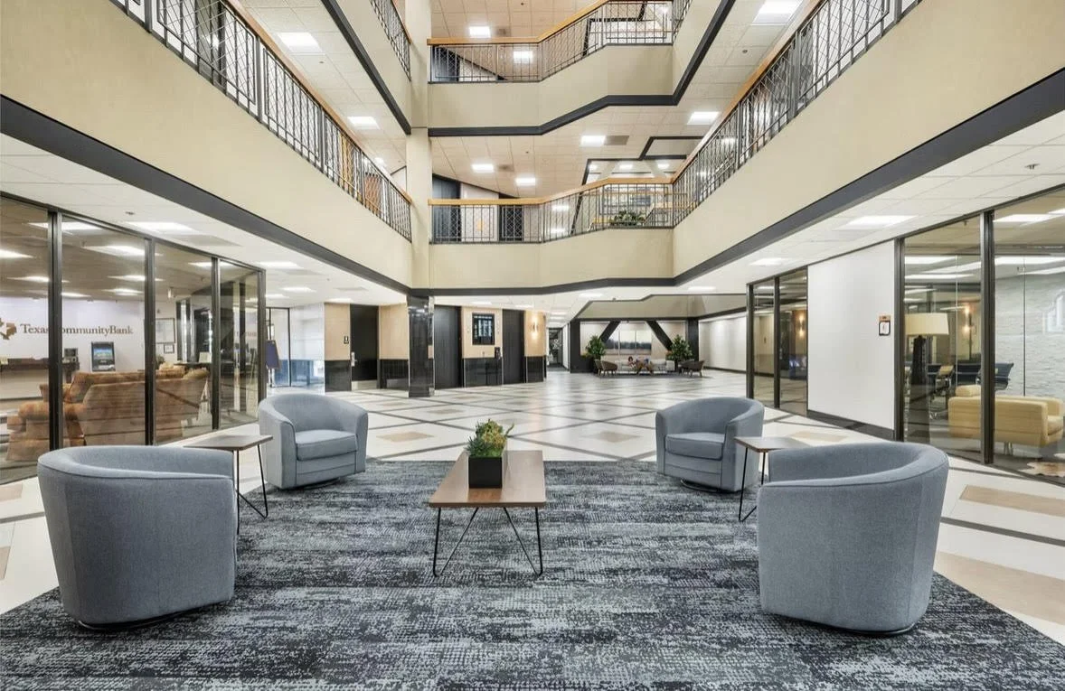 Lobby area with four gray chairs arranged around a coffee table on a dark rug, glass walls showing offices, and a multi-story open atrium with railings and plants.