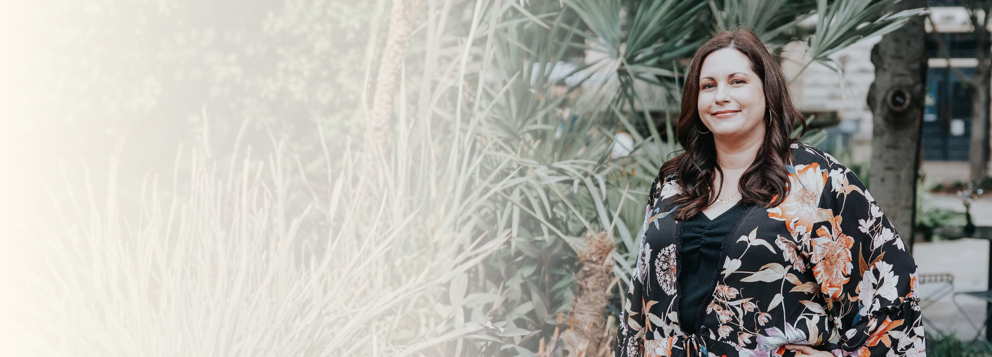 Casey Tully wearing a black floral kimono-style top, standing outdoors in front of green tropical plants, smiling at the camera.