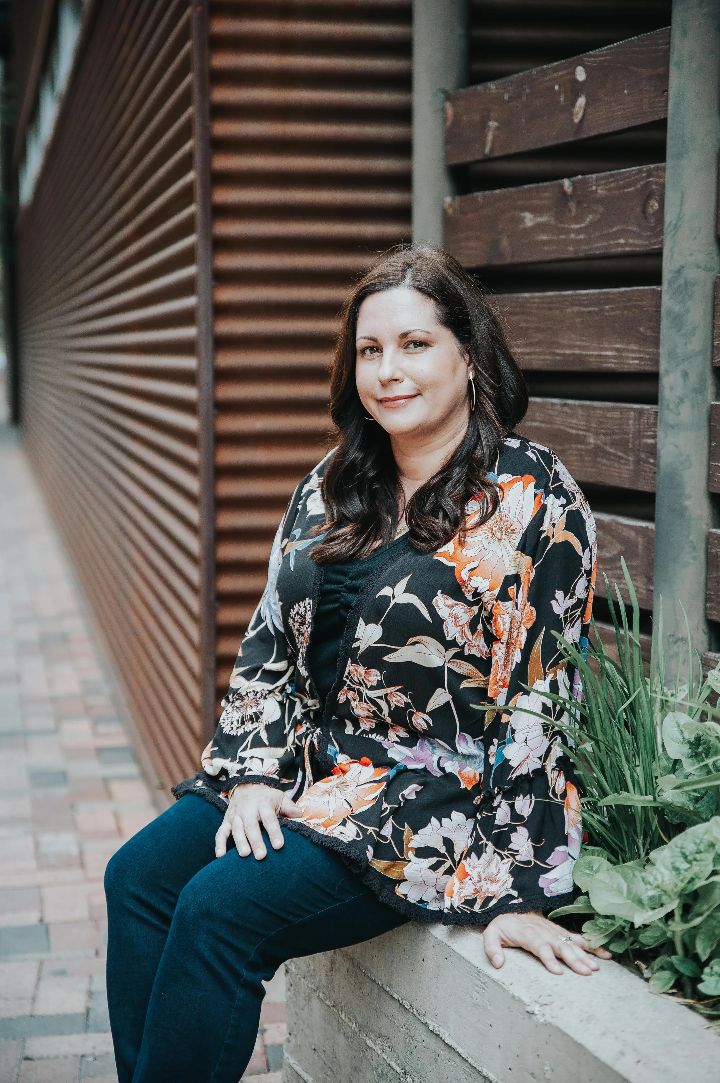 Woman sitting on a concrete ledge outdoors, wearing a floral black top and jeans, with dark hair and hoop earrings, posing confidently near a wooden wall and some greenery.