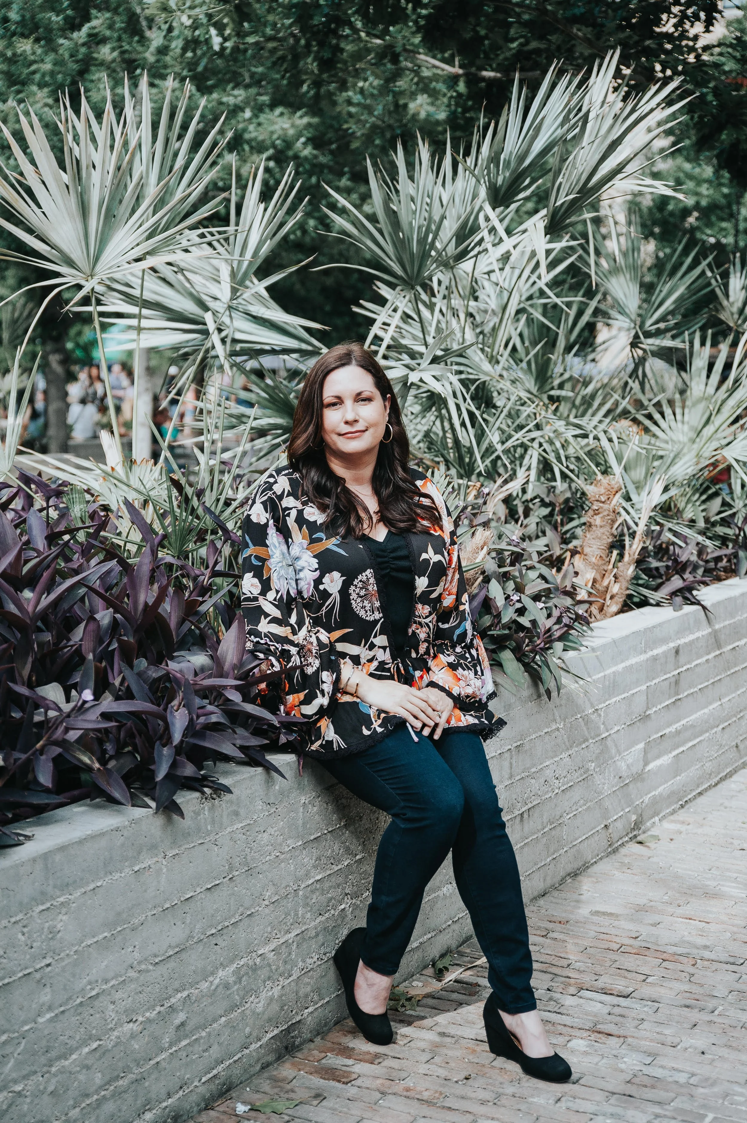 Casey Tully wearing a floral blazer, black top, dark jeans, and black high heels, sitting on a concrete ledge in front of tropical plants.