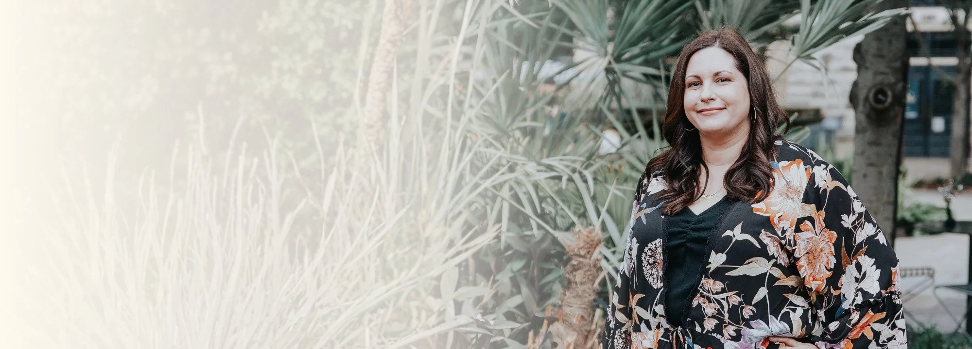 A woman with long dark hair smiling outdoors, wearing a black floral-patterned kimono-style jacket over a black top, with greenery and plants in the background.