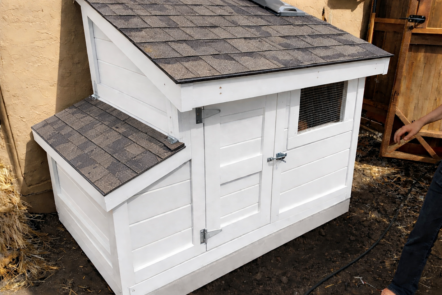 A small white wooden animal shelter with a sloped roof covered in brown shingles, situated outdoors against a yellow wall and a wooden fence.