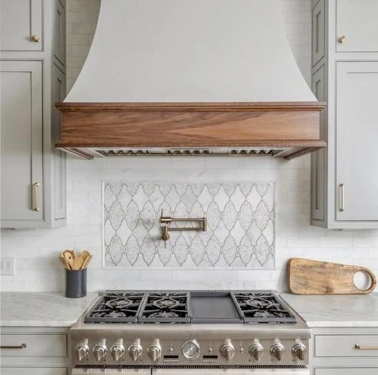 Modern kitchen with gray cabinets, a white tiled backsplash with a decorative pattern, a stainless steel stove, a wooden cutting board, and a black utensil holder on the counter.