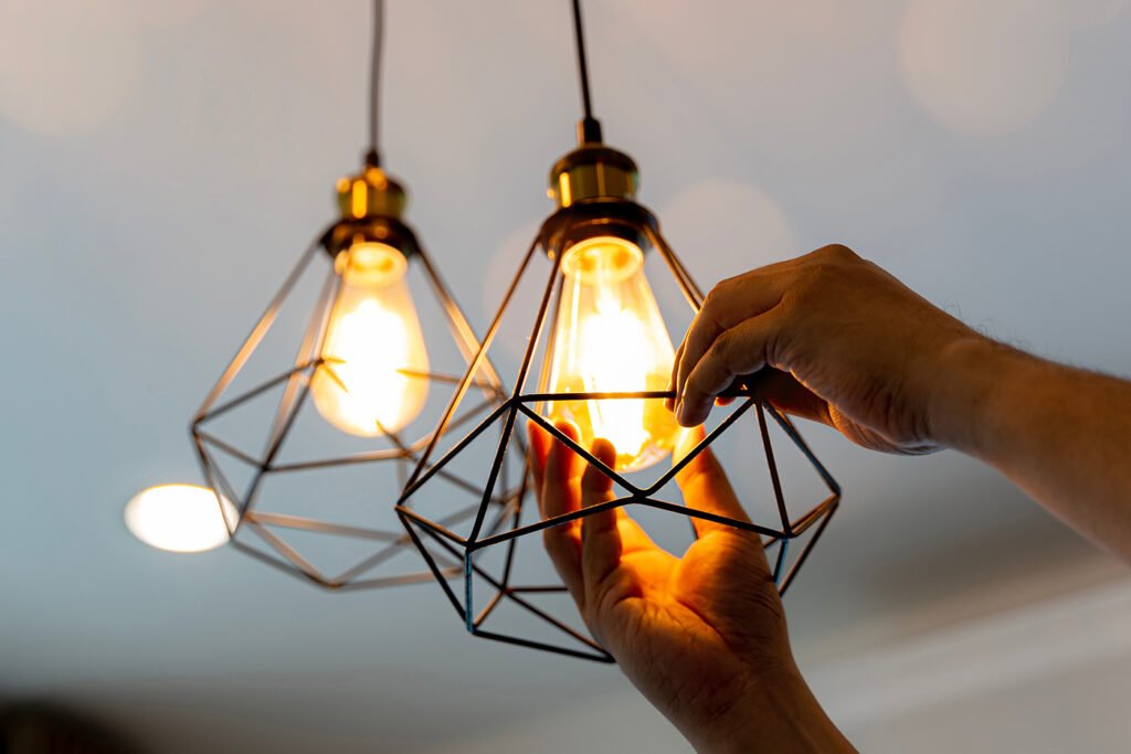 A person adjusting a modern, geometric pendant light fixture with four exposed filament bulbs hanging from a ceiling.