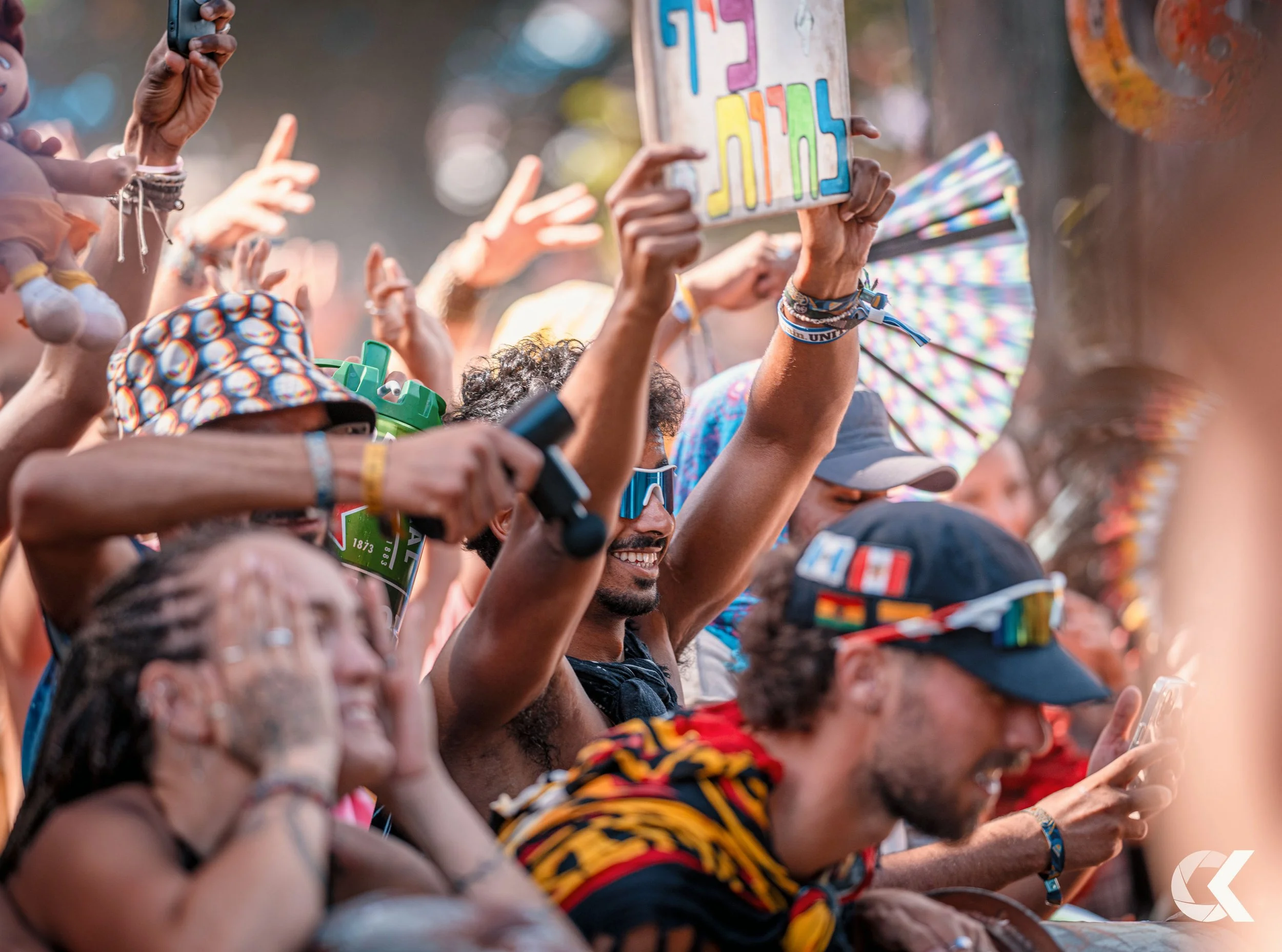 Crowd of diverse people at an outdoor event, some holding signs, wearing sunglasses and colorful clothing, with a joyful atmosphere.