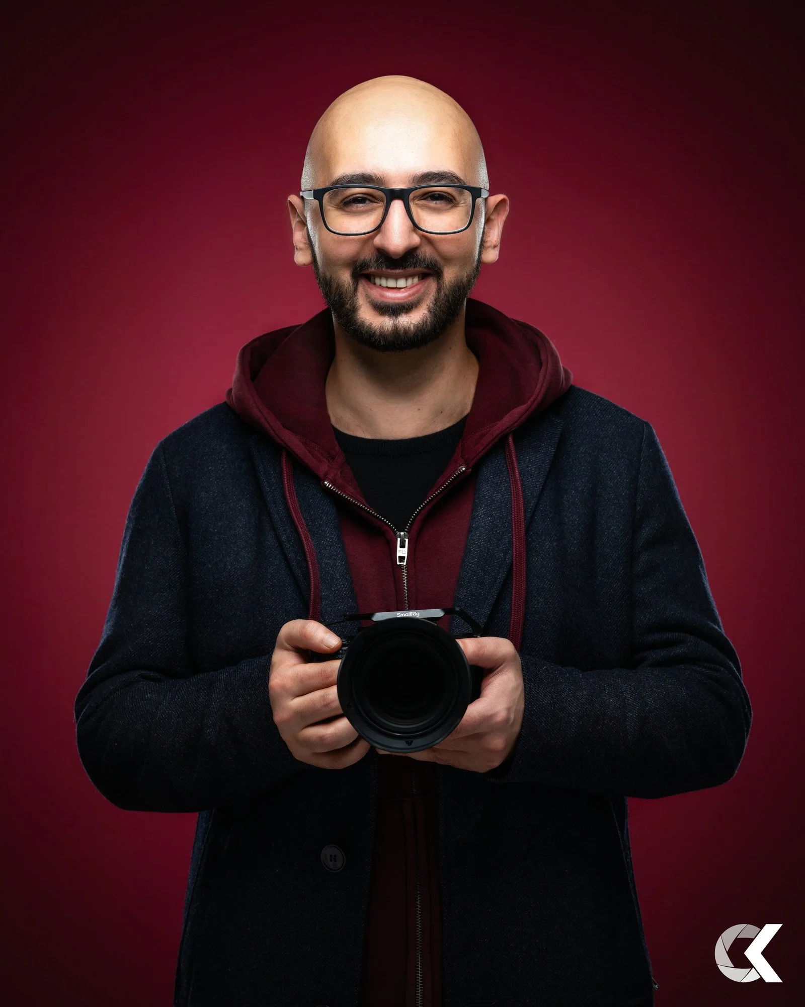 Man with glasses, beard, and shaved head holding a camera, smiling, against a red background.