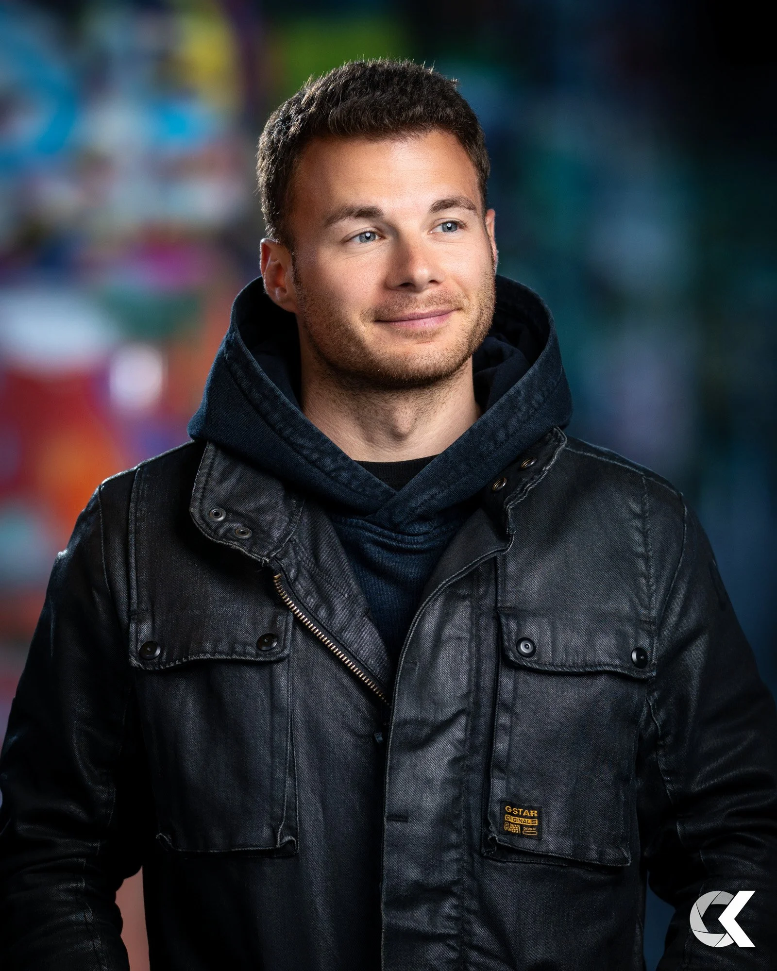 A young man with short brown hair and a light beard, smiling slightly, wearing a black leather jacket over a dark blue hoodie, standing outdoors at night with a blurred colorful background.