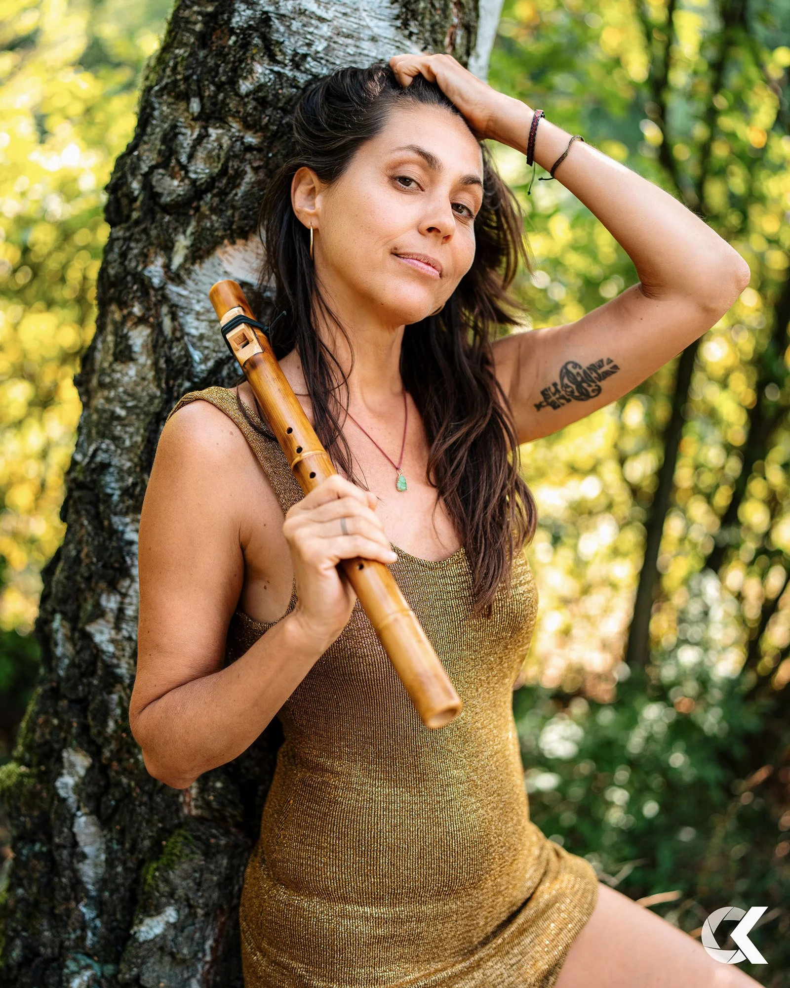 A woman with long dark hair, wearing a sleeveless gold dress, holding a wooden flute, standing outdoors against a tree trunk in a forest with green leaves.