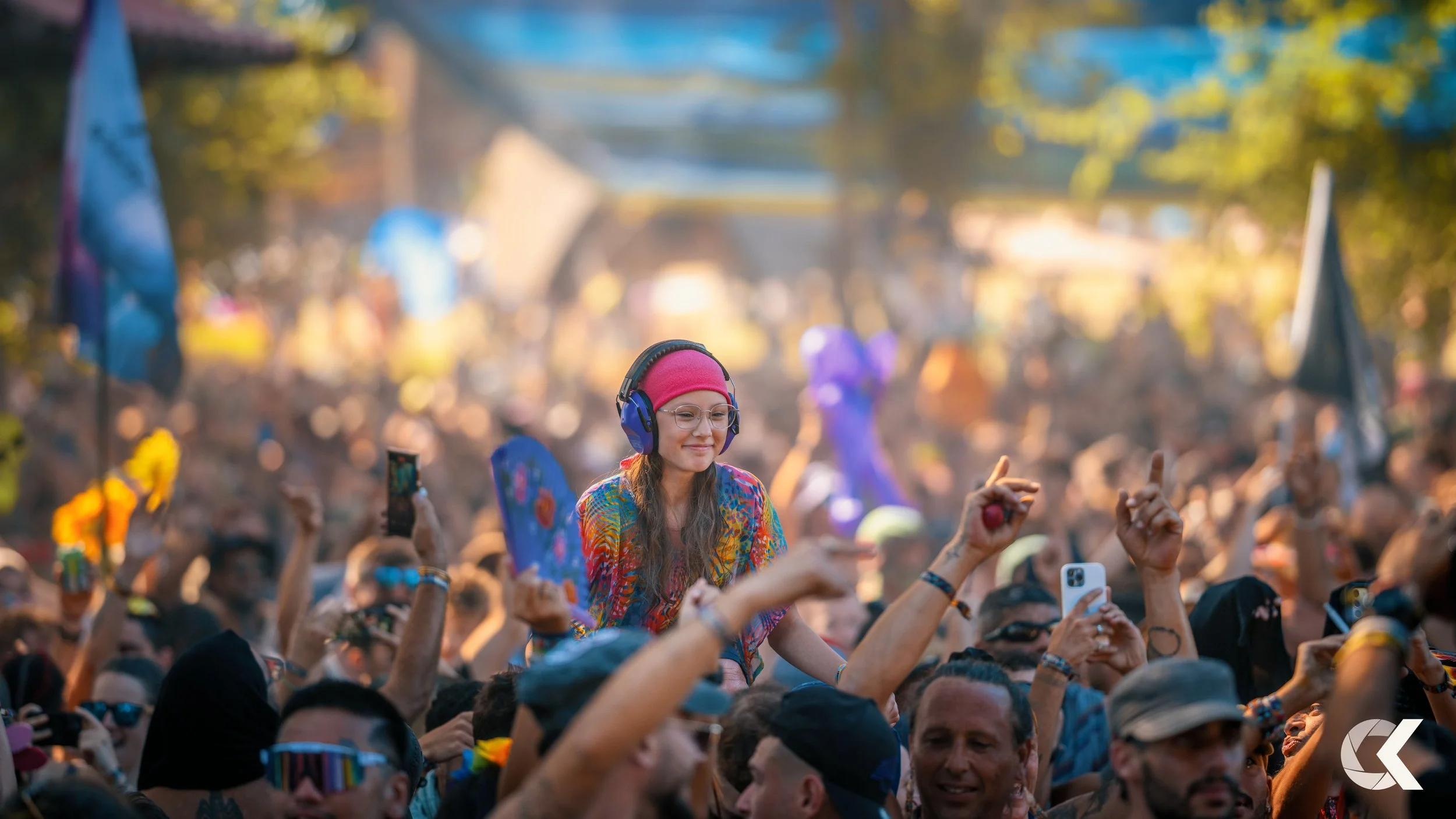 Crowd of people at an outdoor music festival, with a woman in a pink headband and large headphones being lifted above others, many with phones taking pictures or videos, colorful flags, and trees in the background.