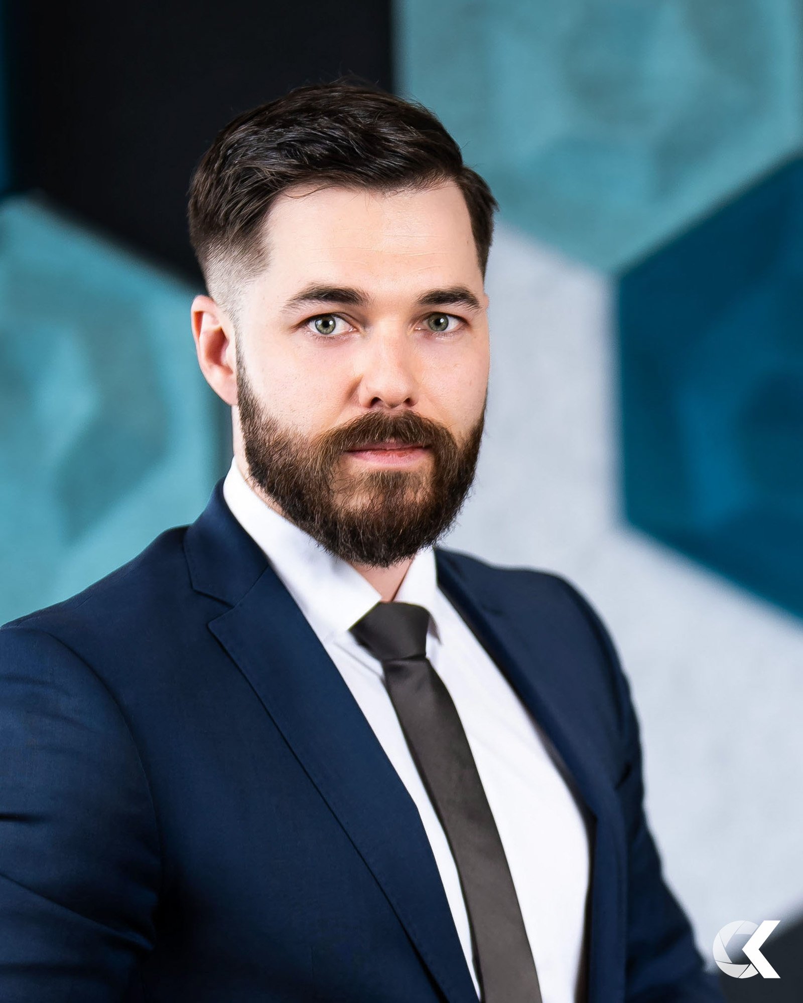 A man with a beard and short dark hair dressed in a navy blue suit, white shirt, and black tie, looking at the camera with a serious expression, against a geometric background with shades of blue.