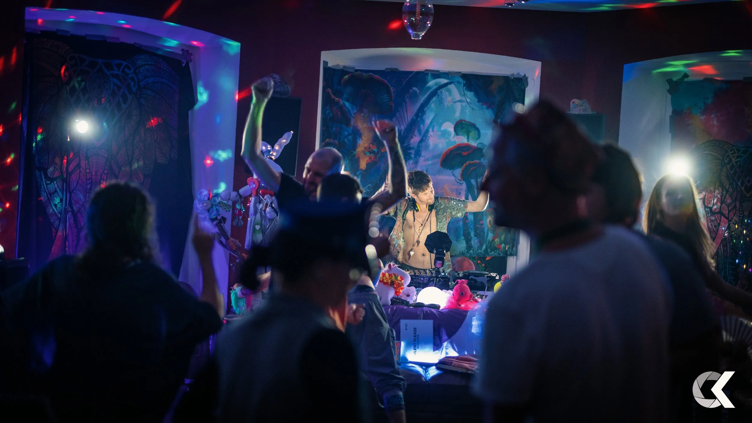 People dancing in a dark, colorful room with neon lights, DJ on stage, and vibrant artwork in the background.
