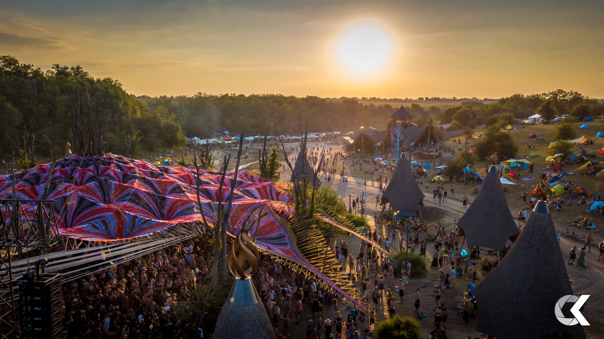 Sunset over a crowded outdoor festival with tents, structures, and people enjoying activities.