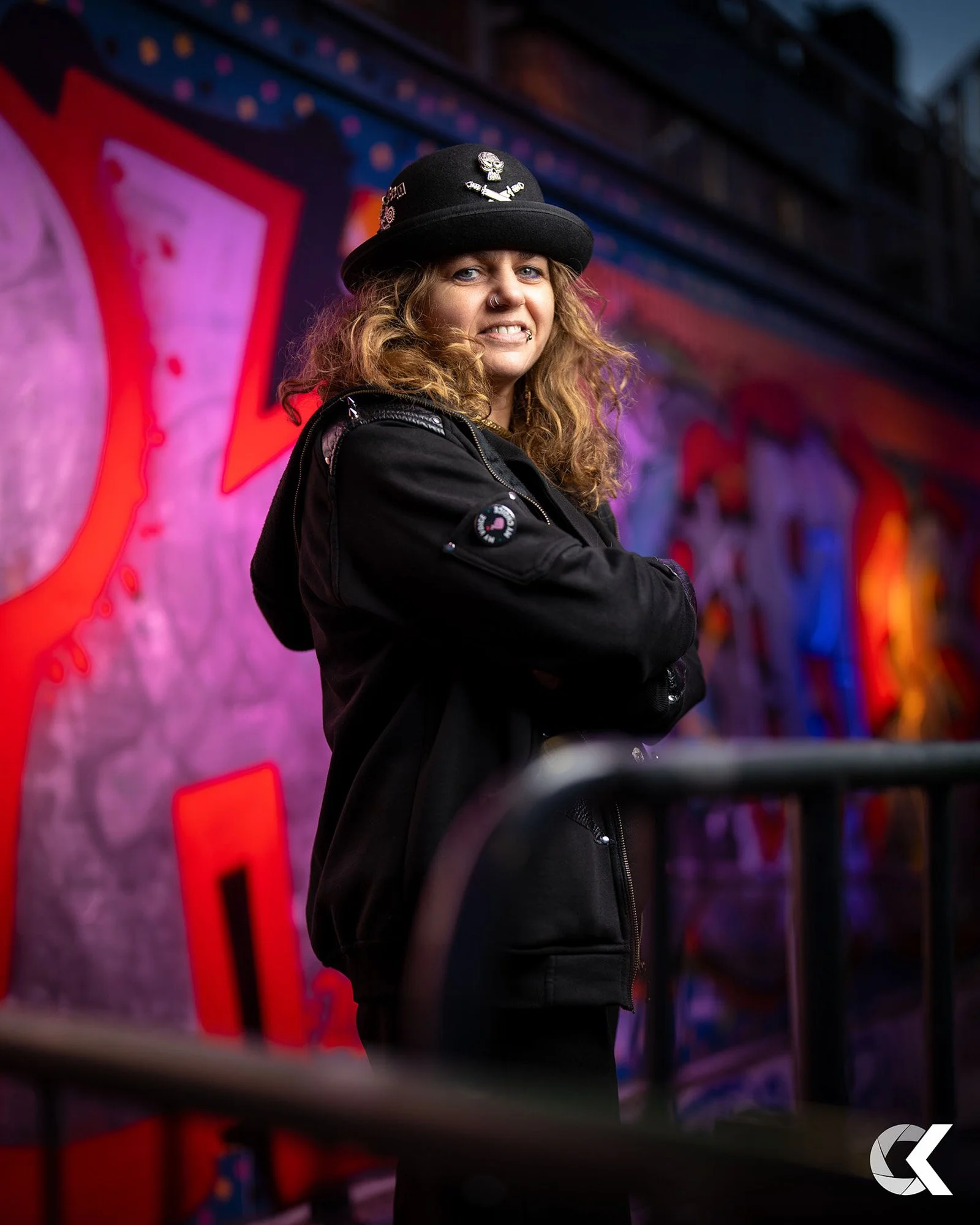 A woman with curly hair and a nose ring stands with her arms crossed in front of a colorful graffiti wall, wearing a black jacket and a black hat with patches, smiling slightly.