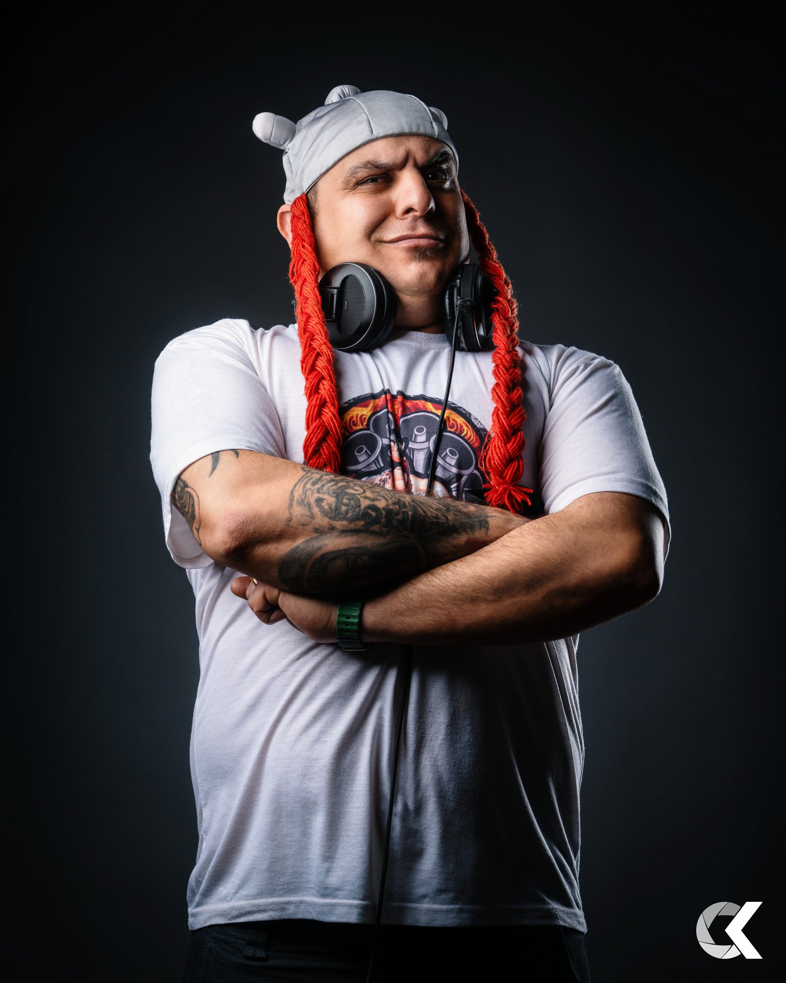 A man with tattoos wearing a gray bear-themed beanie with red long braids, headphones around his neck, and a white T-shirt with a colorful graphic, standing against a dark background.