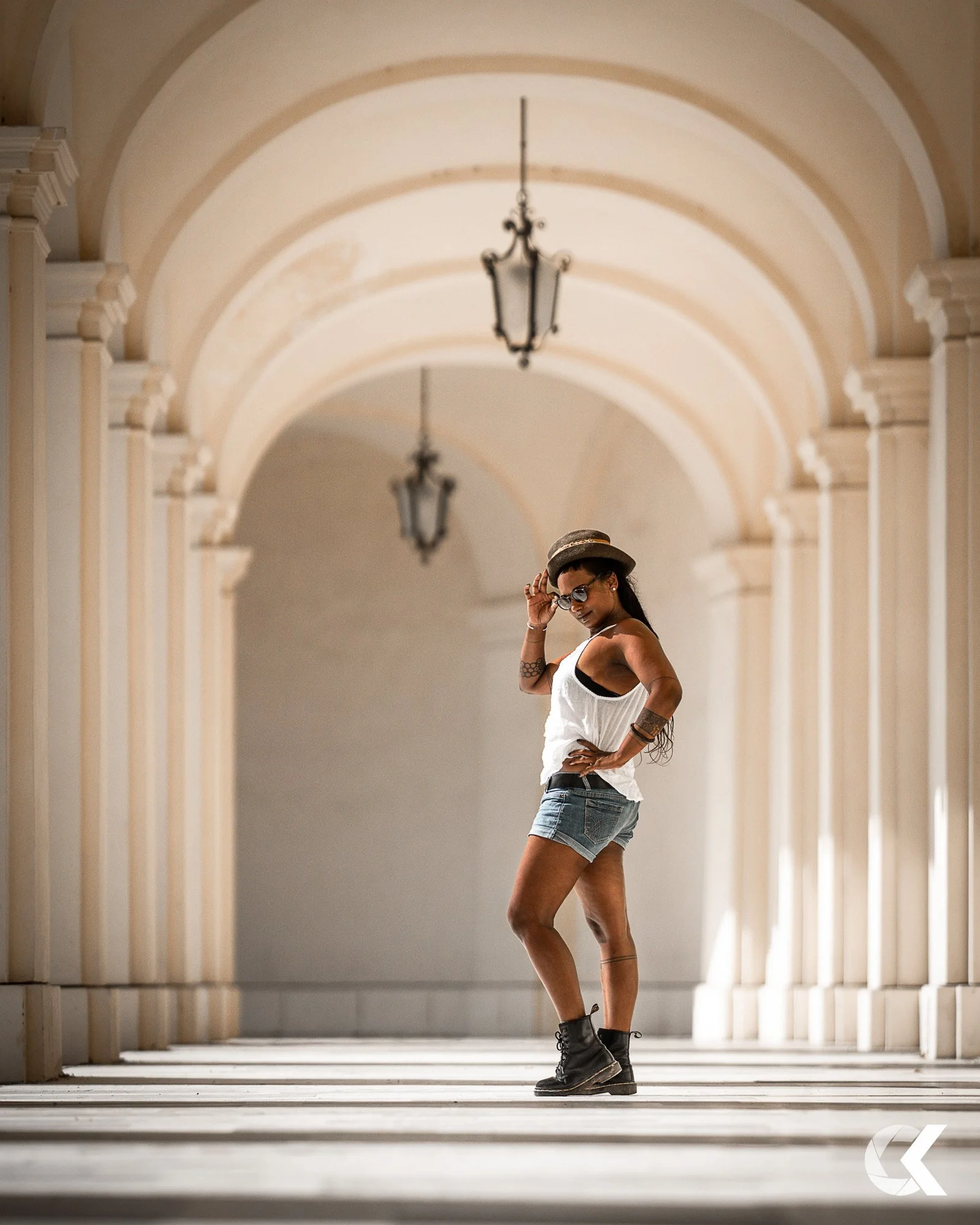 A woman in casual attire, including a white top, denim shorts, black boots, sunglasses, and a fedora hat, stands confidently in a covered walkway with arched ceilings and hanging lanterns.