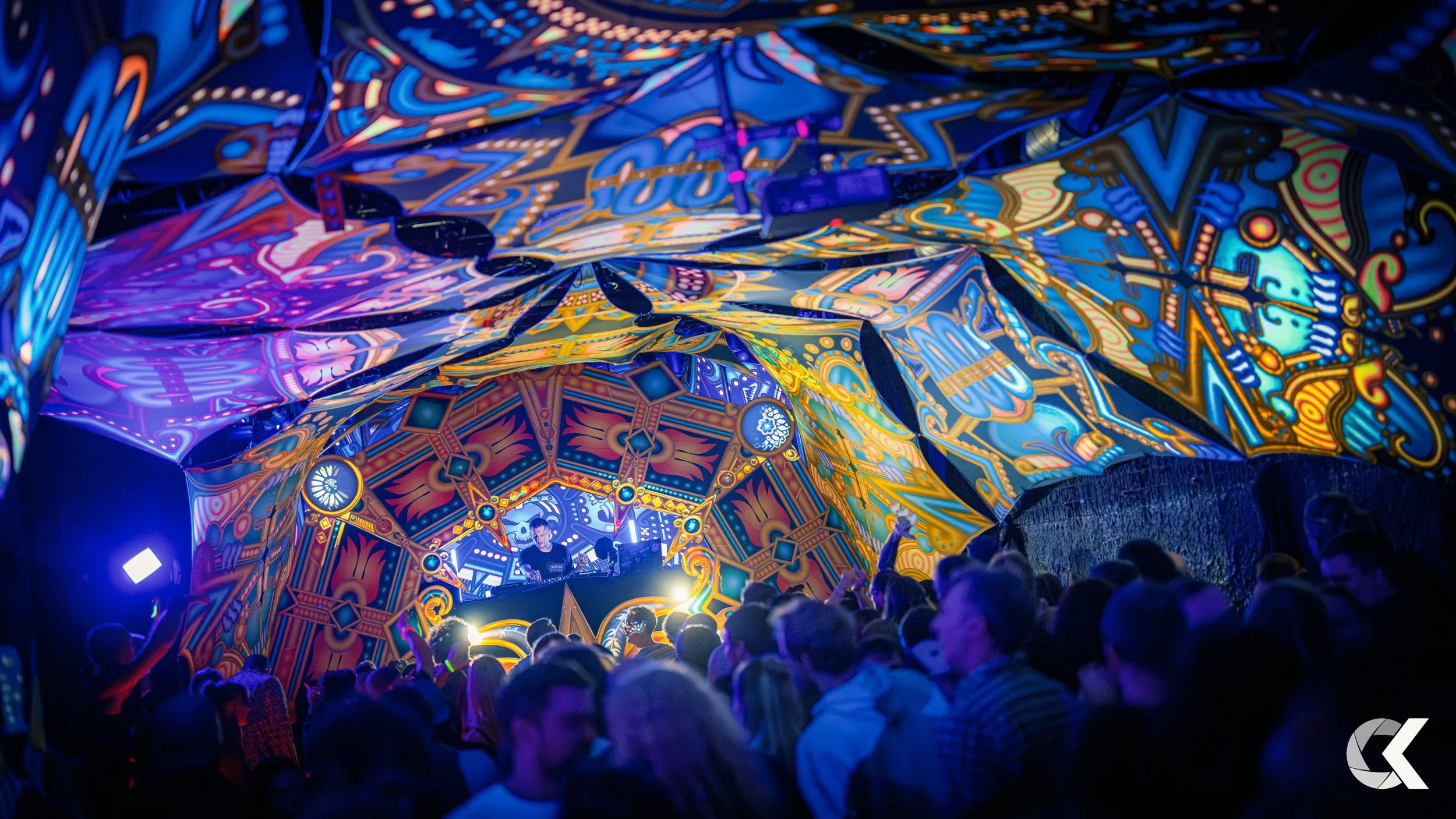 A crowded concert with a colorful, abstract, painted ceiling and a DJ performing on stage.