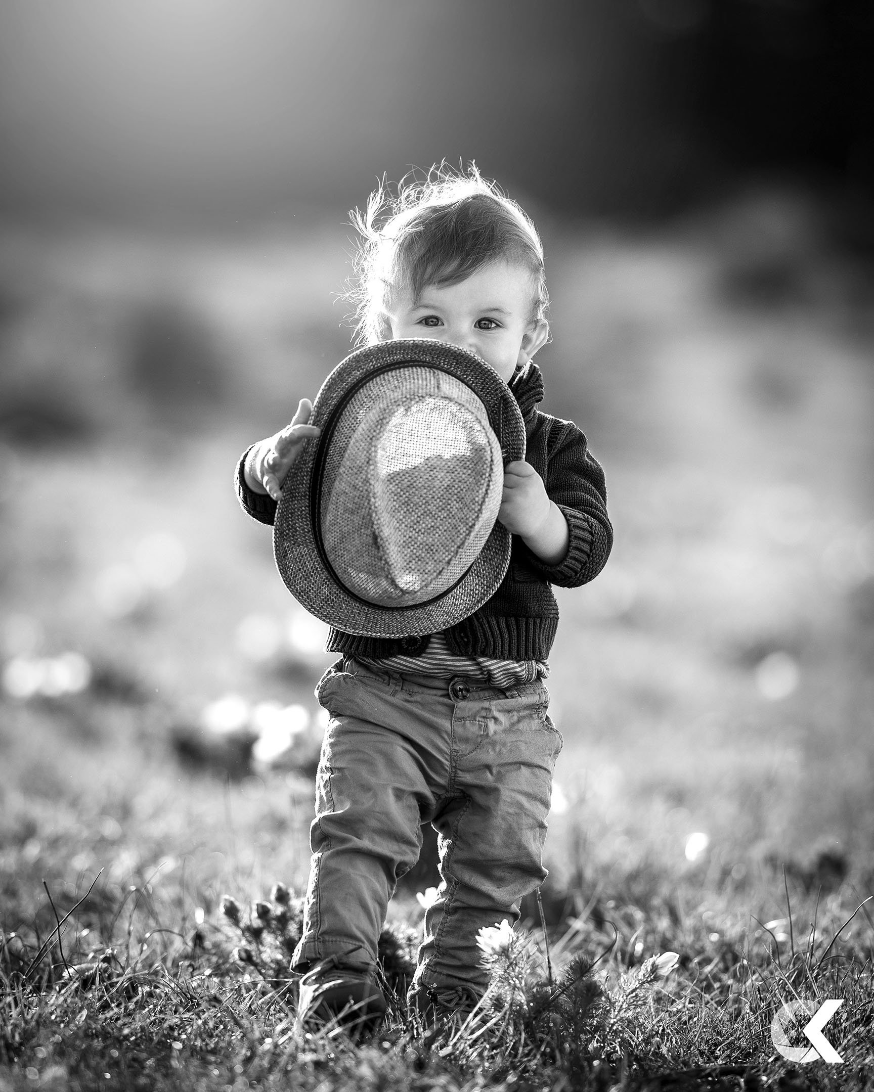 A black and white photo of a young child standing outdoors on grass, holding a hat with a mountain scene on it, and looking at the camera.