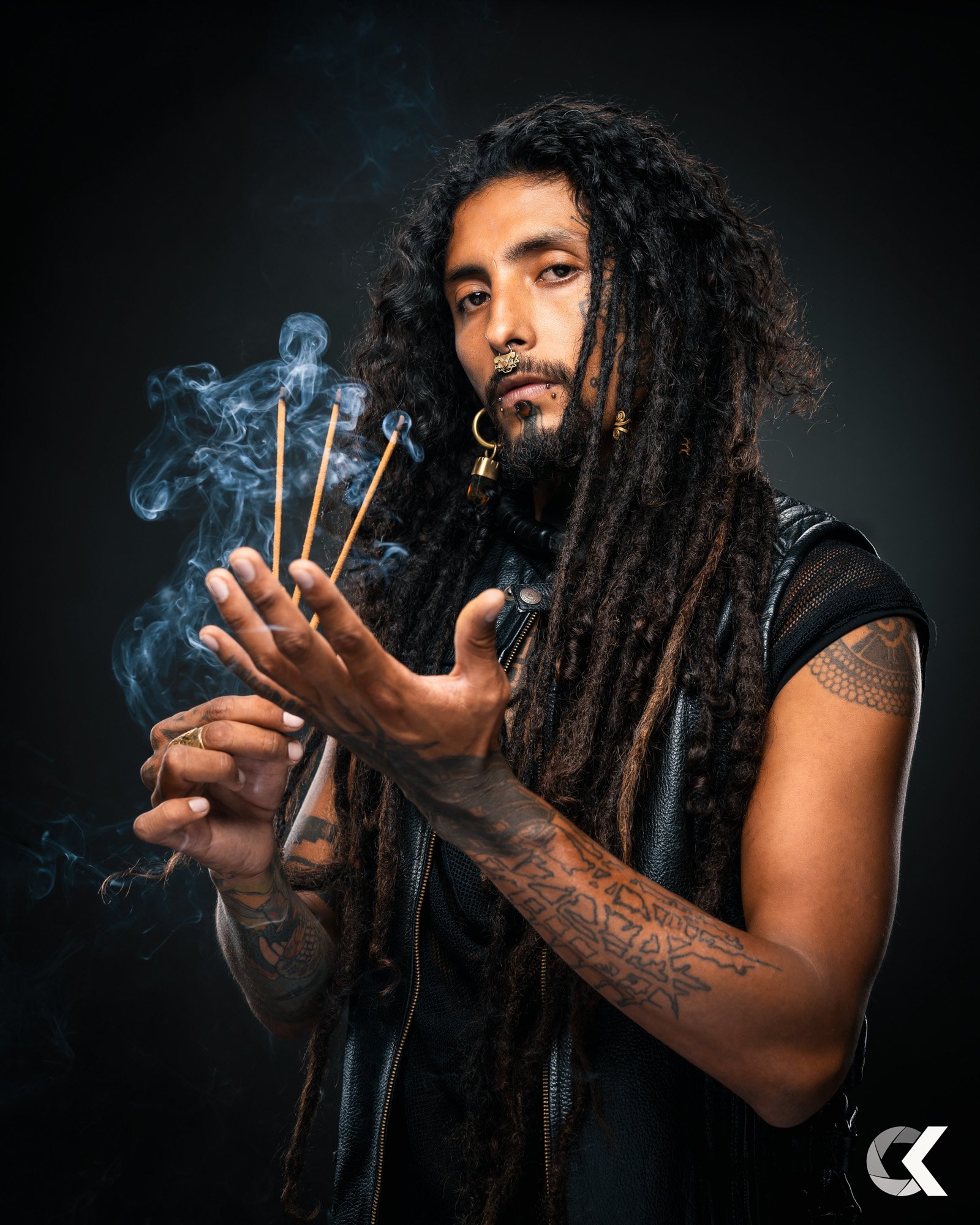 A person with long dreadlocks, tattoos, and multiple facial piercings holds incense sticks with smoke rising, against a dark background.
