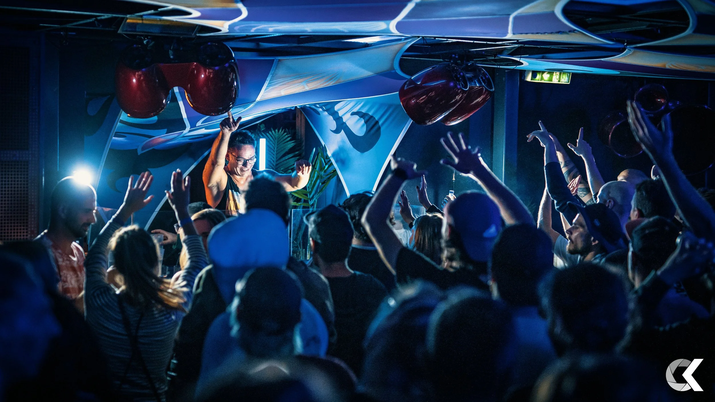Crowd dancing at a nightclub with a DJ on stage, blue lighting, and balloons hanging from the ceiling.
