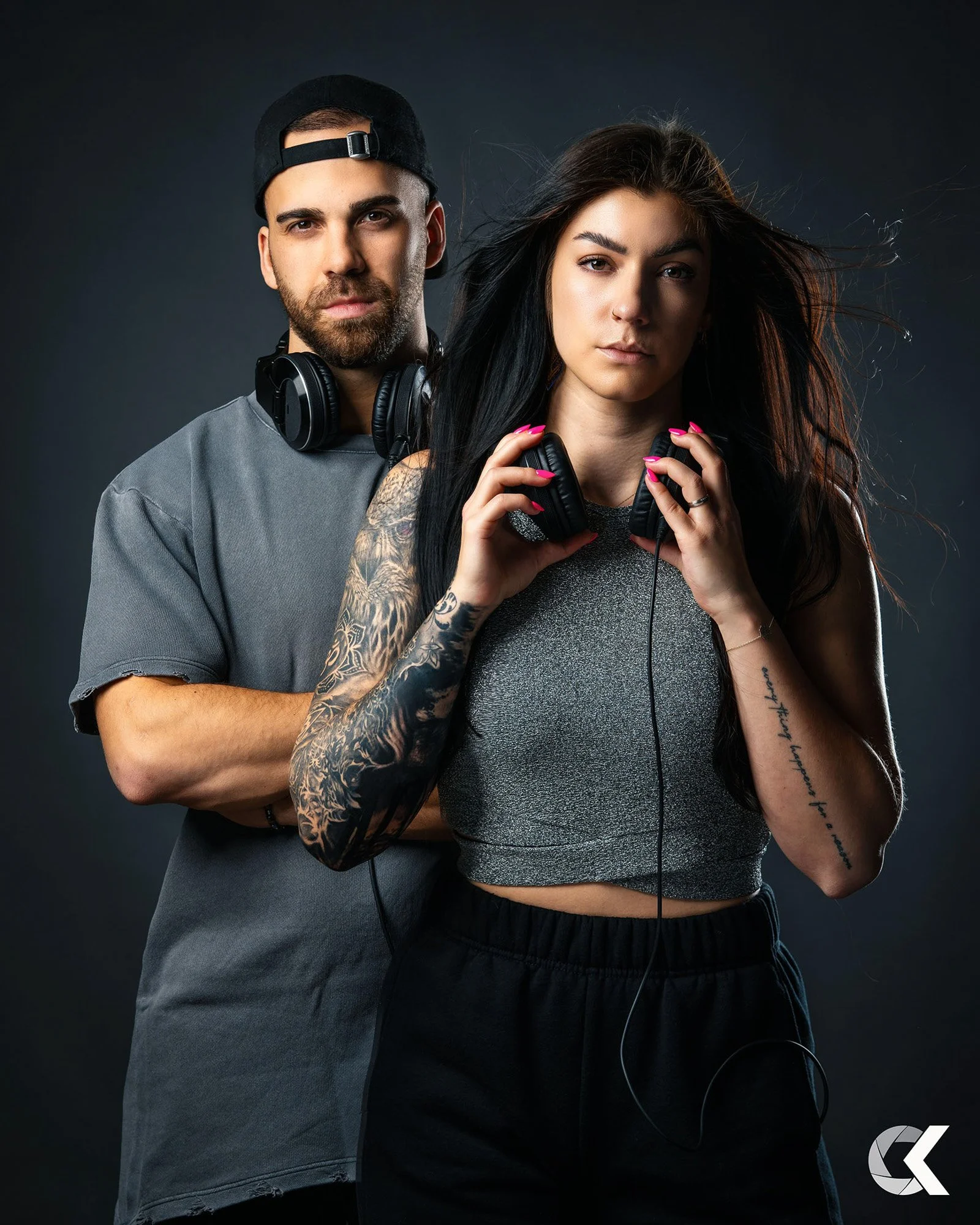 A young man and woman posing with headphones against a dark background. The man has a beard, wears a backward cap, and gray t-shirt. The woman has long black hair, tattoos on her arms, and wears a gray crop top and black pants. They look directly at 