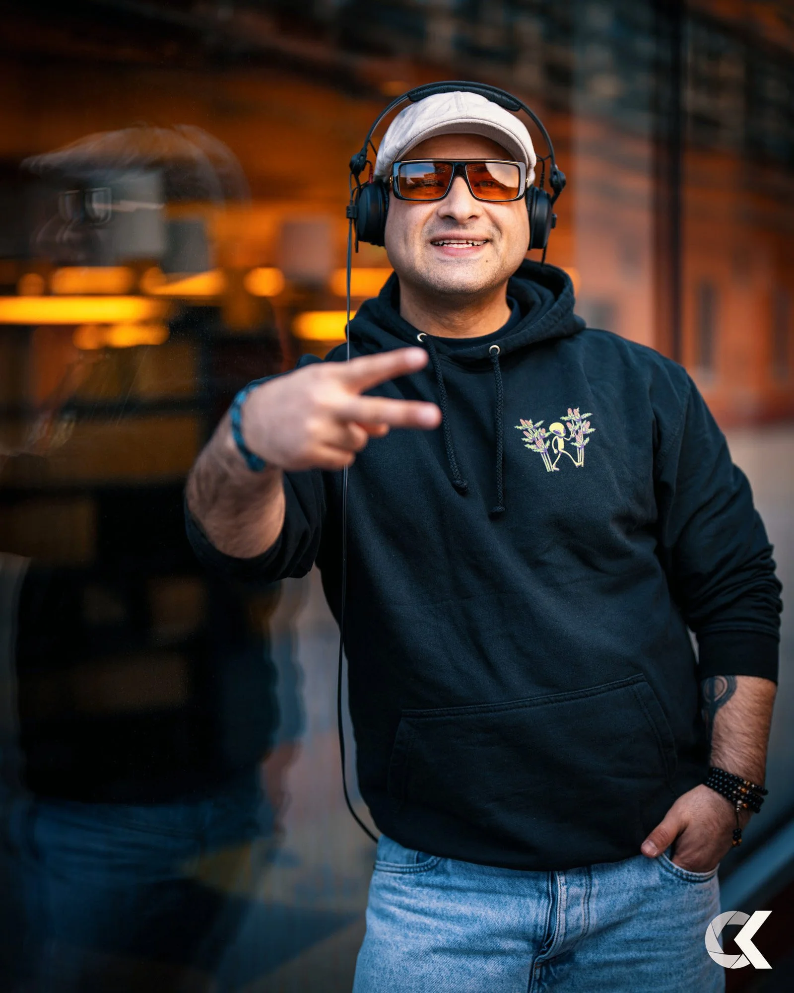 A man wearing sunglasses, a hoodie, and a cap, smiling and flashing a peace sign in front of a reflective glass window, outdoors during dusk.