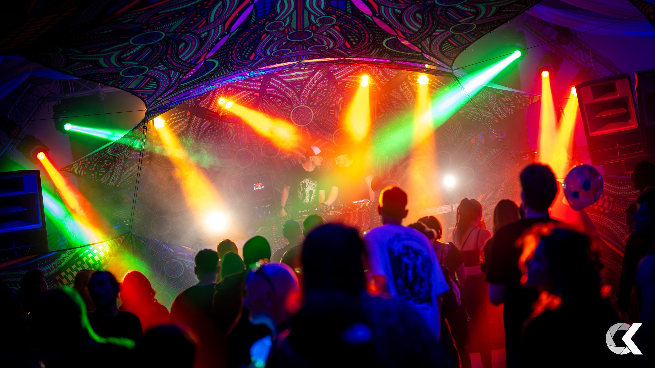 A nightclub with colorful lights and a DJ performing on stage surrounded by a crowd of people dancing.