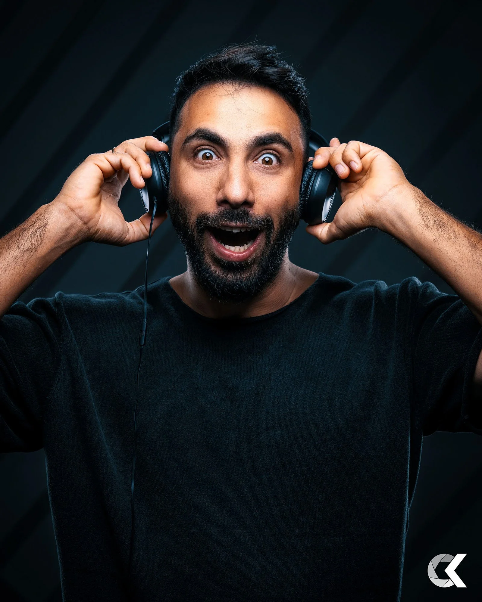 A man with dark hair and a beard wearing a black shirt, holding headphones to his ears, smiling with an open mouth, in a dark setting.