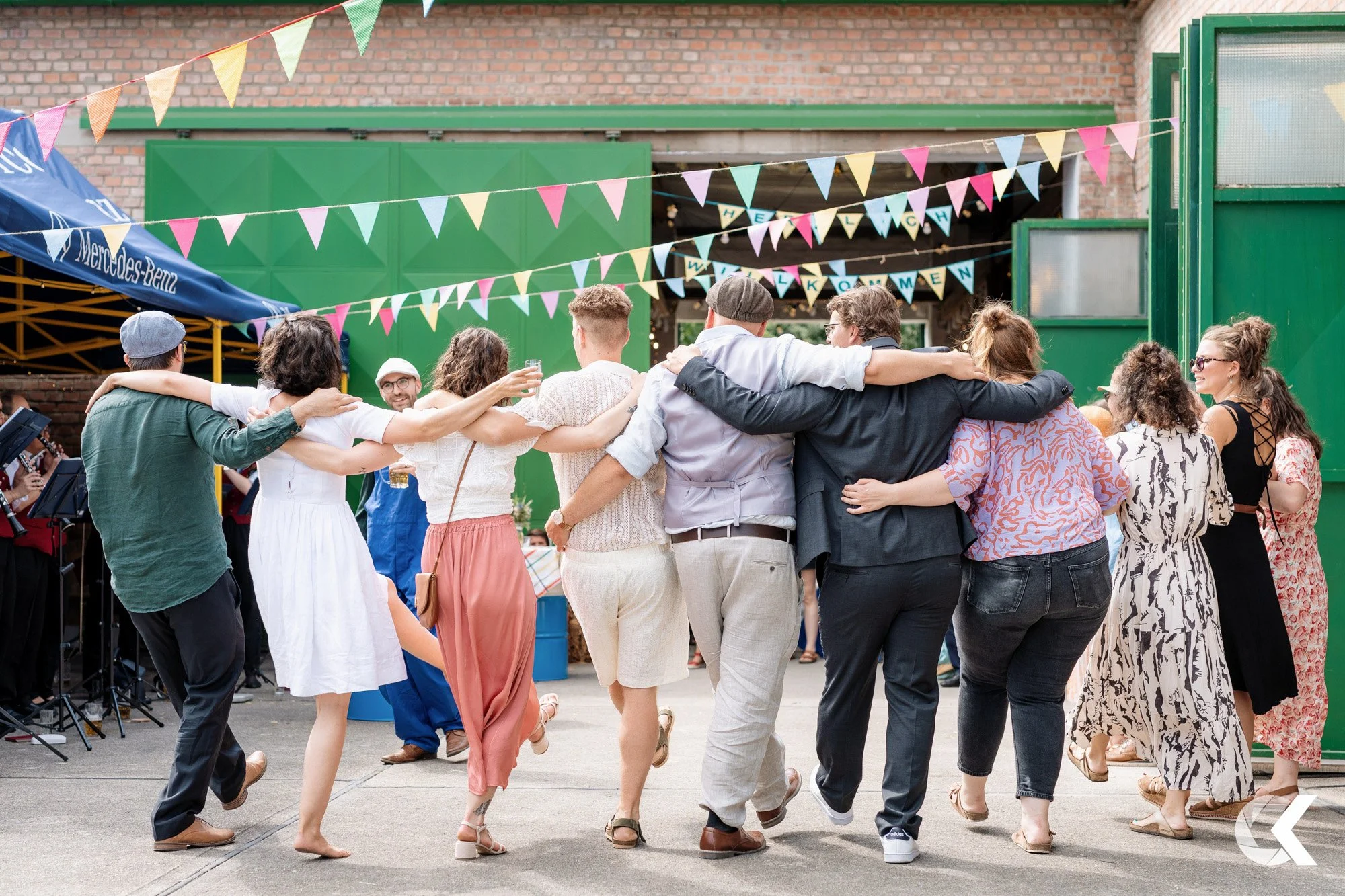 Group of people dancing in a circle at an outdoor party, with colorful bunting hanging overhead.