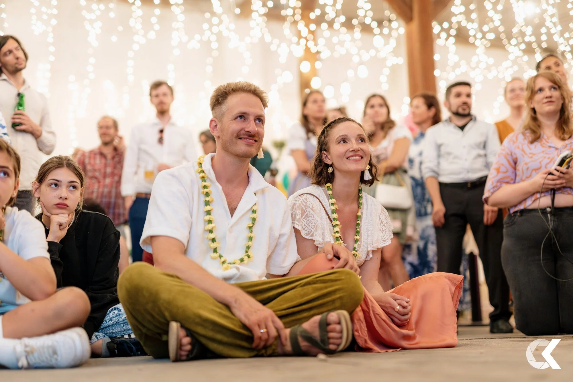 People sitting on the floor at a celebration event, wearing traditional leis, with string lights hanging above in a decorated indoor setting.