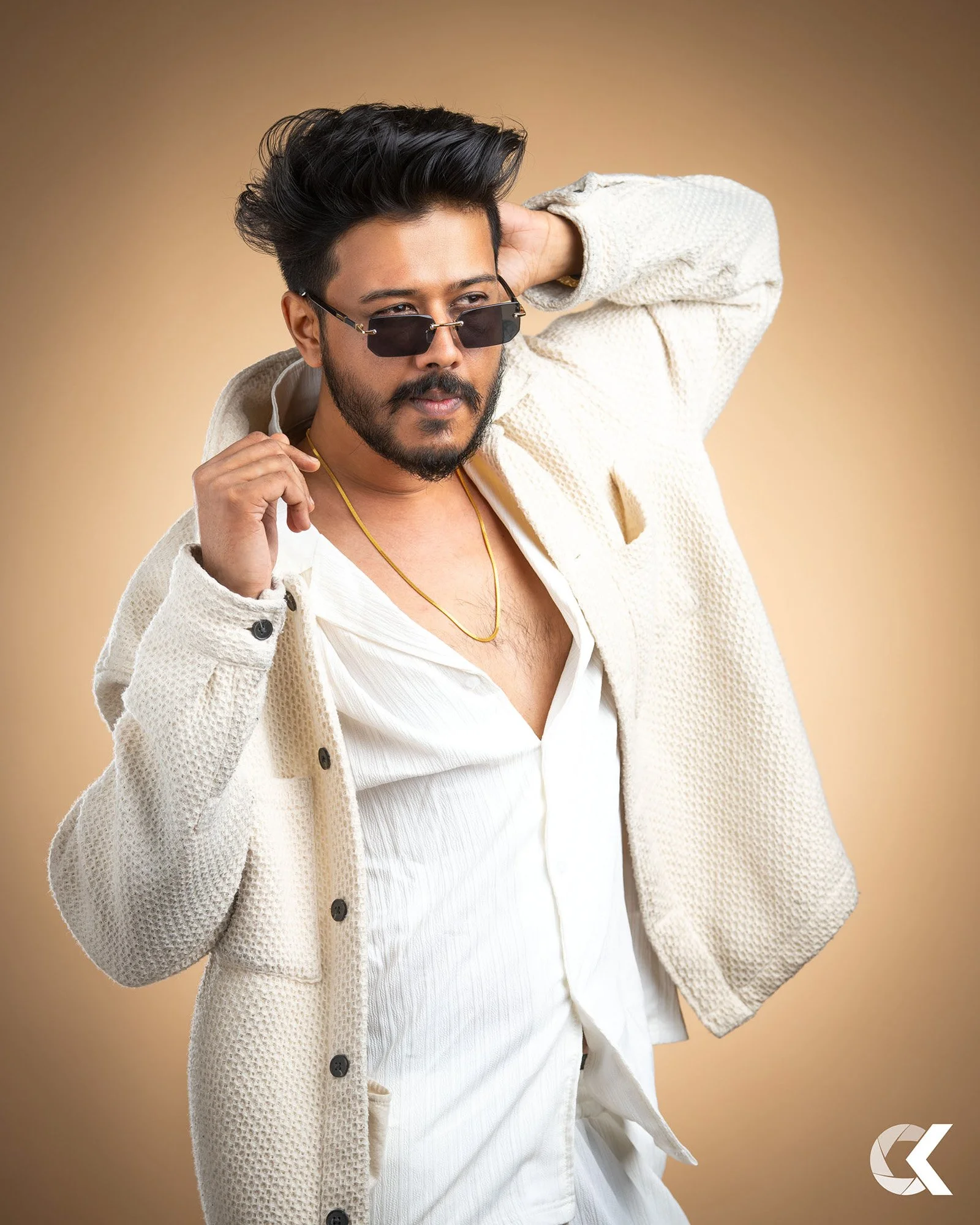 Ein Mann mit dunklem Haar, Bart und Sonnenbrille trägt eine cremefarbene Jacke, ein weißes Hemd und eine goldene Halskette. Er posiert vor einem beigen Hintergrund.