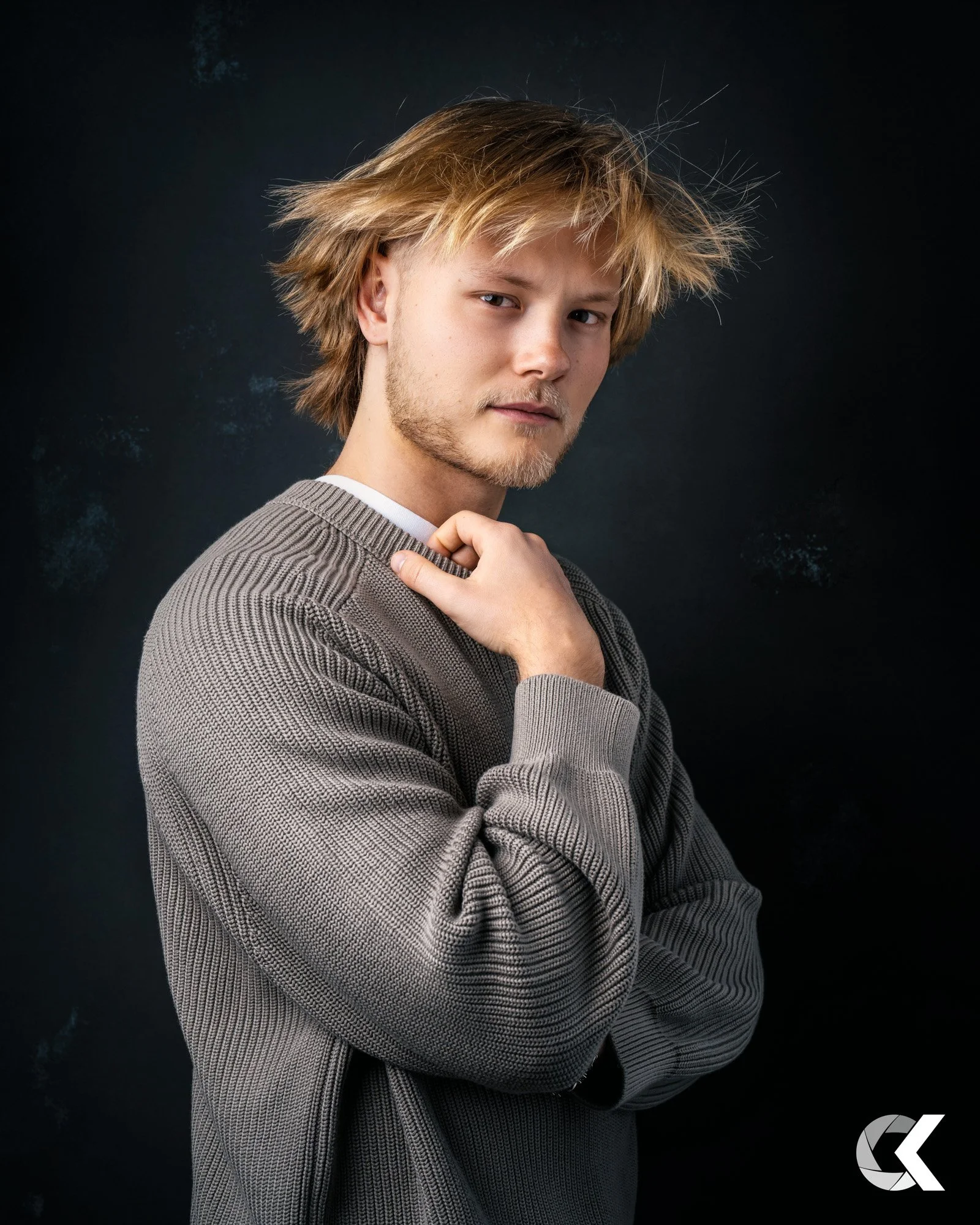 Junger Mann mit Blond, zerzaustem Haar, futuristische Kleidung, betrachtet nachdenklich die Kamera, dunkler Hintergrund.