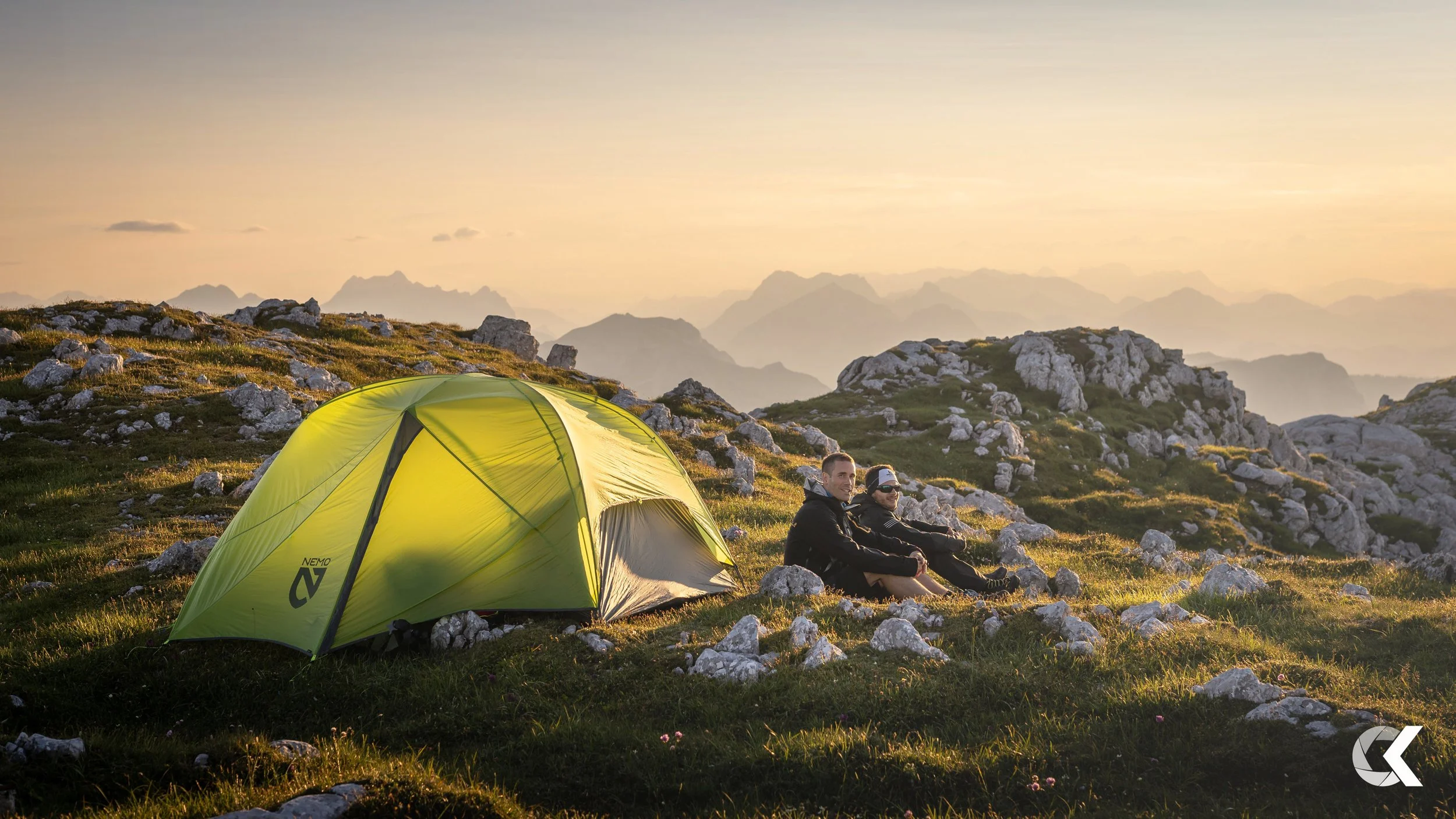 Two people sitting near a yellow tent on a grassy, rocky mountain landscape during sunset, with mountain ranges in the background.