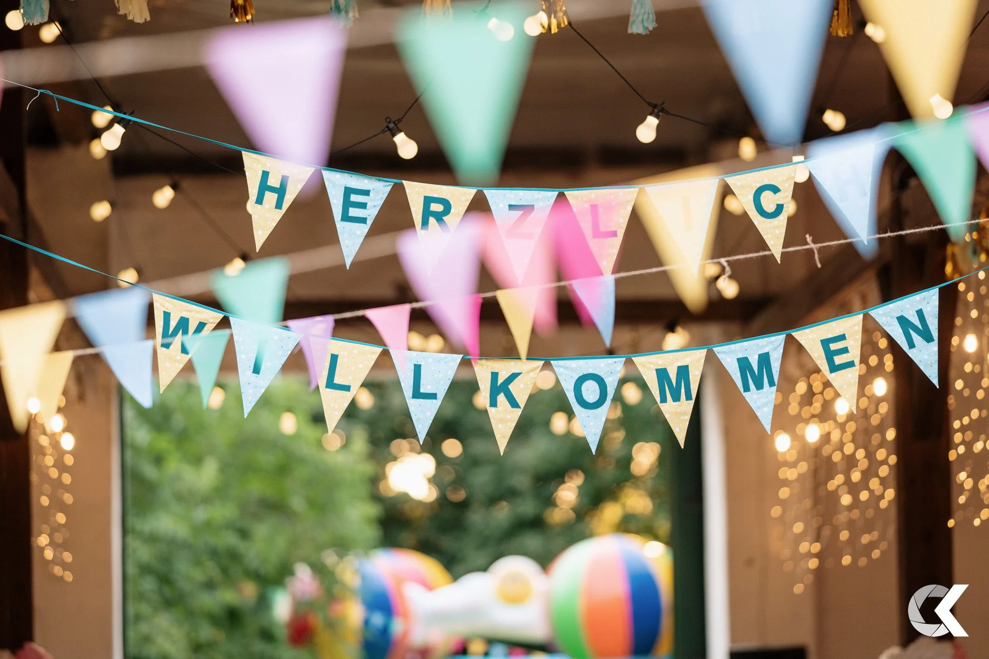 Colorful 'Welcome' and 'Hello' banners hanging indoors with string lights and a blurred outdoor background with trees and balloons.