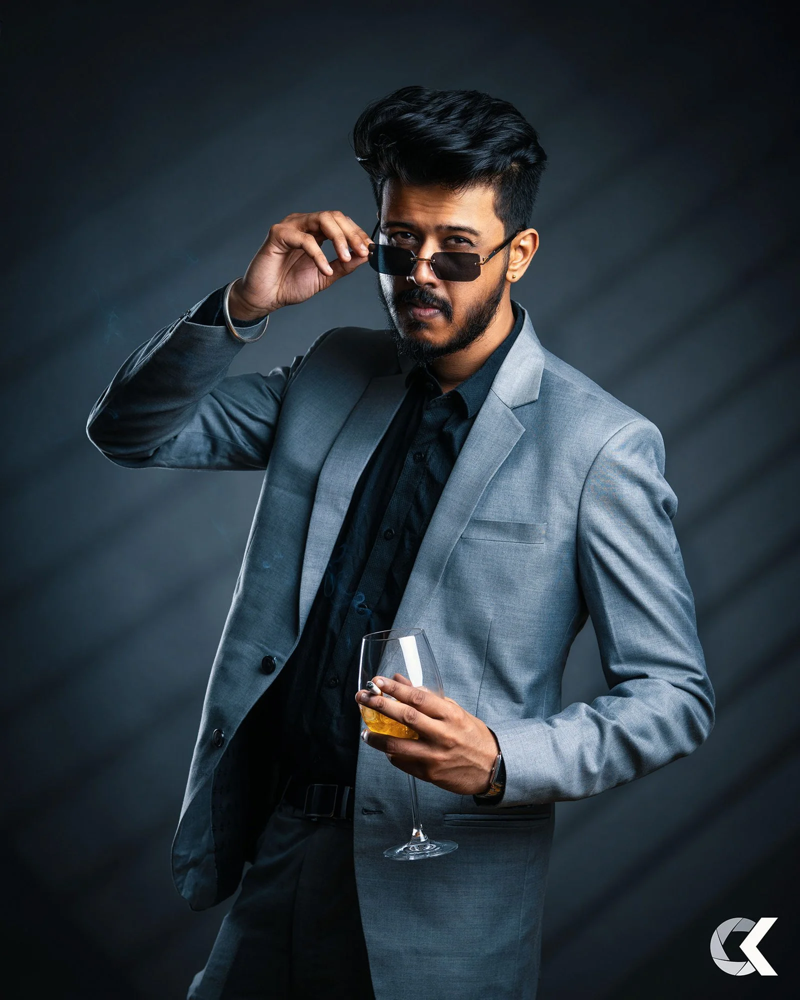 Ein Mann trägt einen grauen Anzug, schwarze Hemd, Sonnenbrille, hält ein Whiskyglas und schaut selbstbewusst in die Kamera.