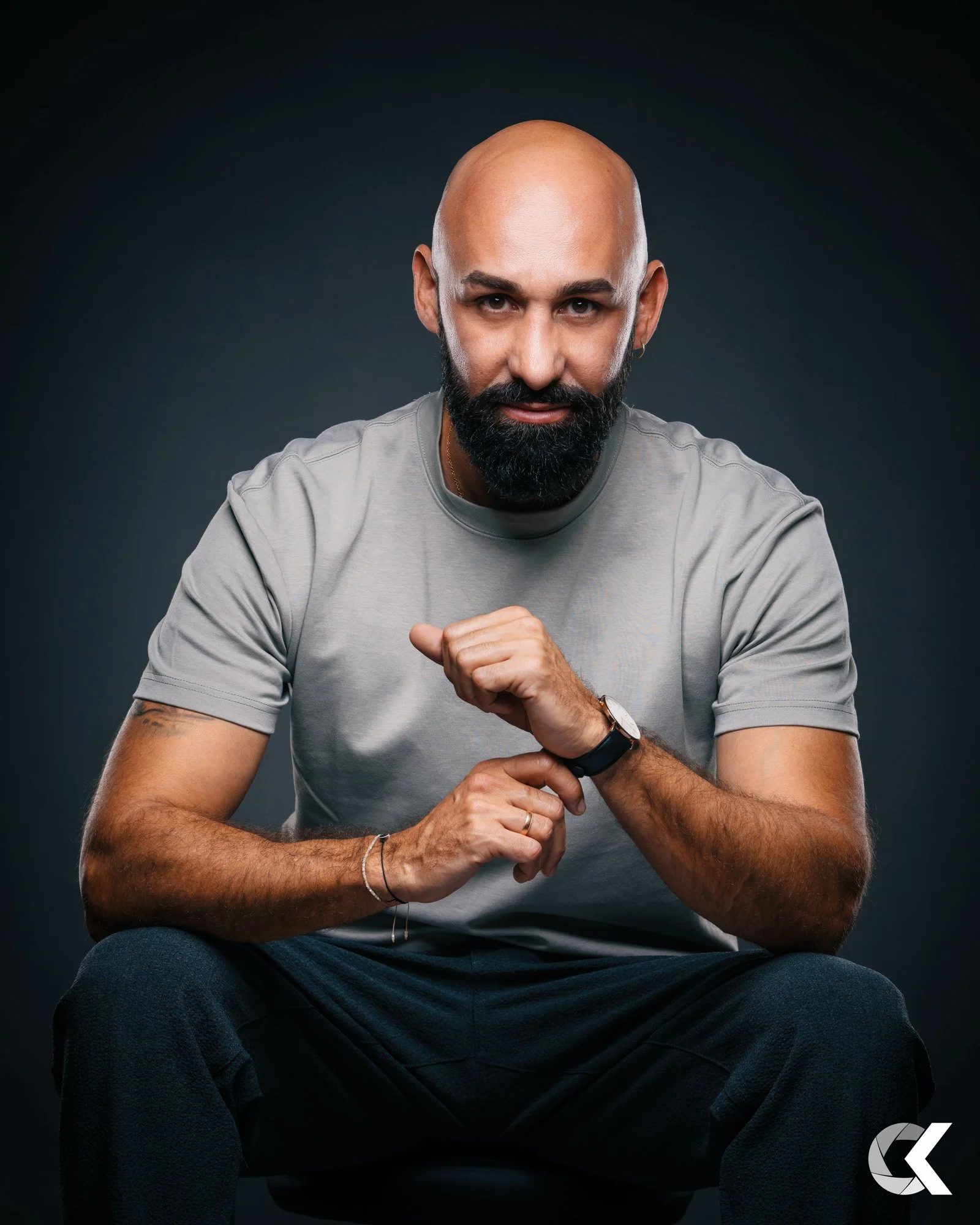 A bald man with a beard looking at the camera, sitting down, wearing a gray t-shirt, and showing off his wristwatch.