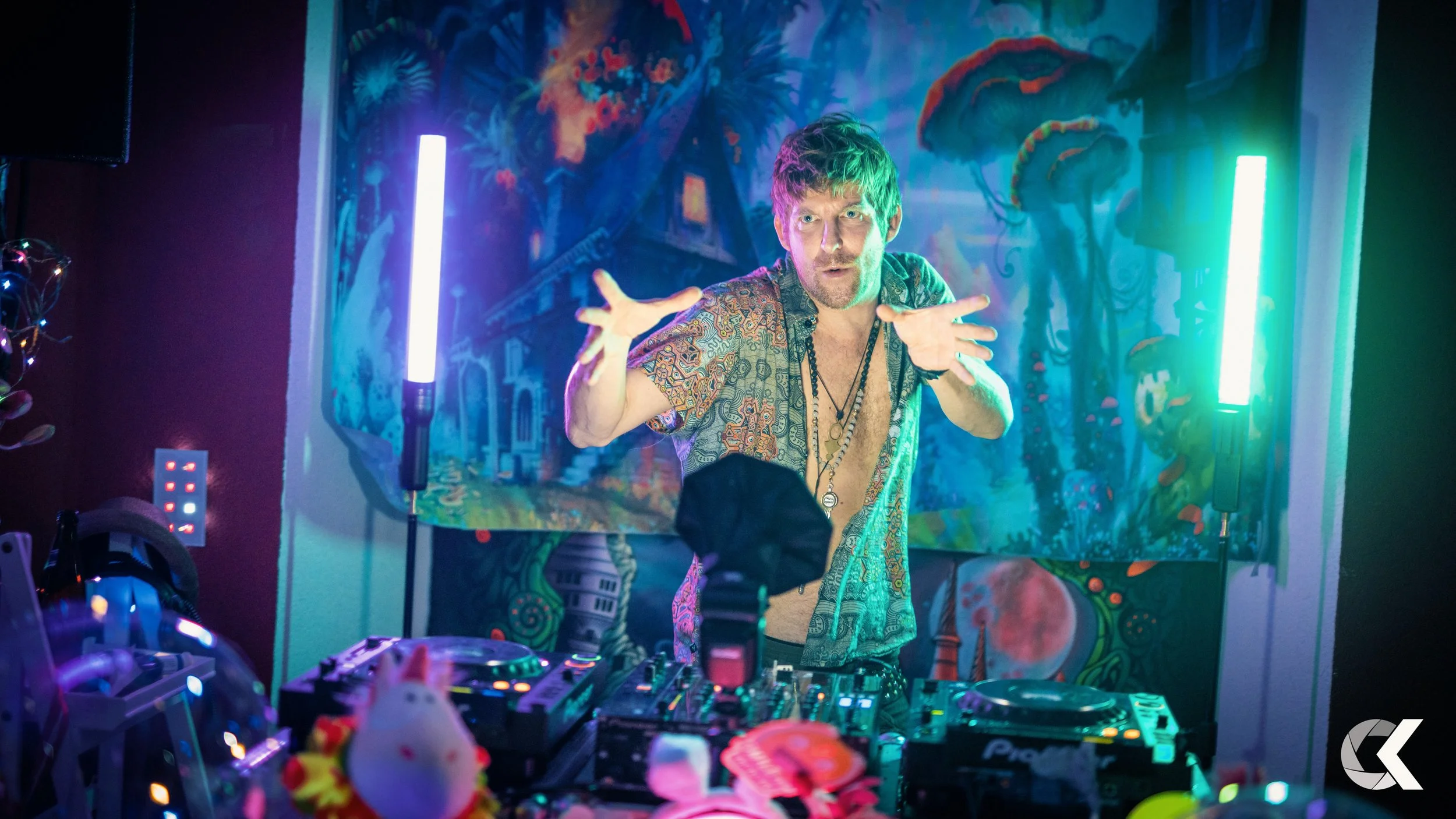 A man with short hair wearing a patterned shirt and necklaces, standing behind DJ equipment, gesturing with raised hands, with a colorful, neon-lit background of a mural of a house and trees.