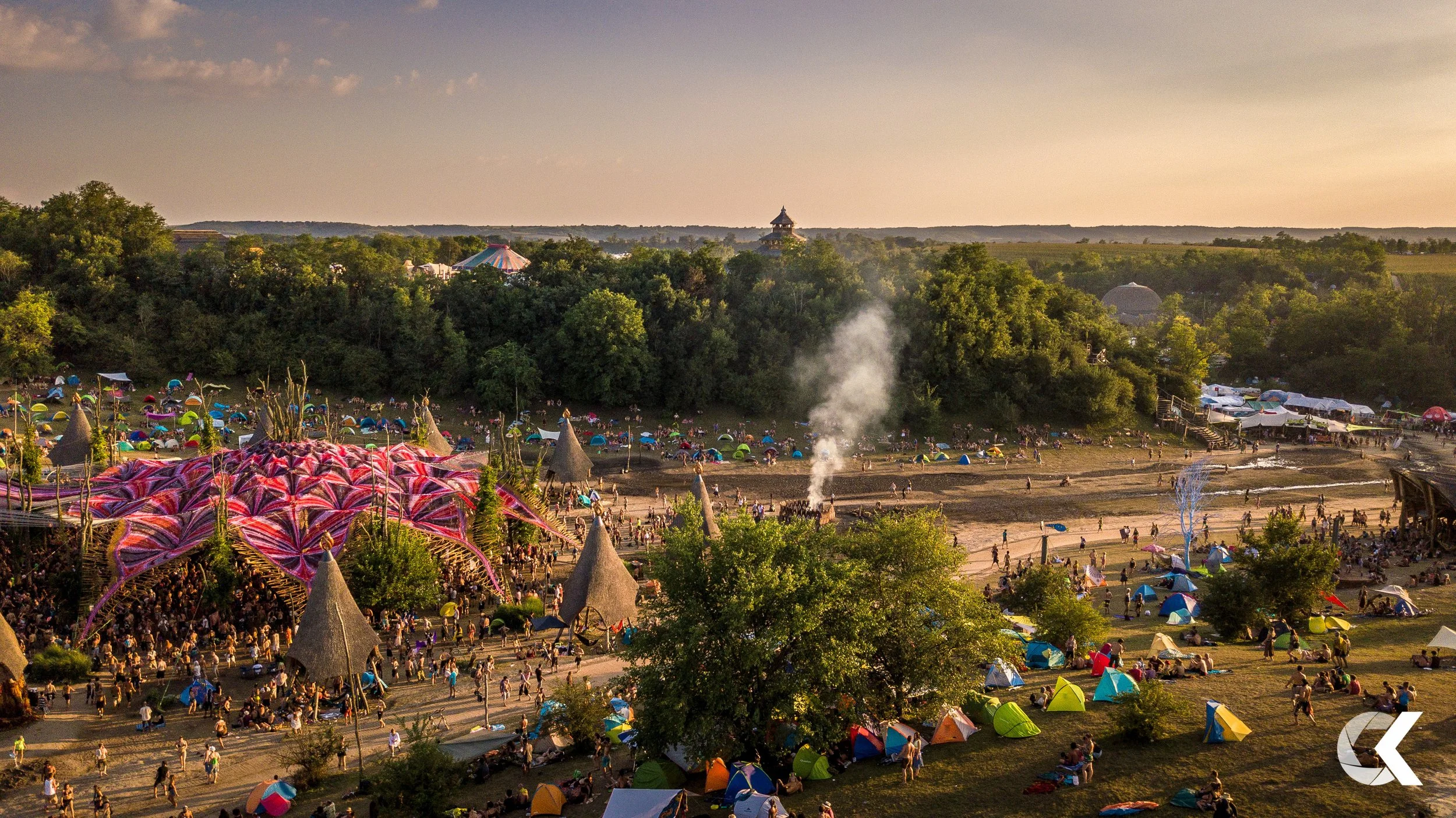 A large outdoor festival scene at sunset with many tents, a decorated stage or structure, and people enjoying various activities in a park surrounded by trees.