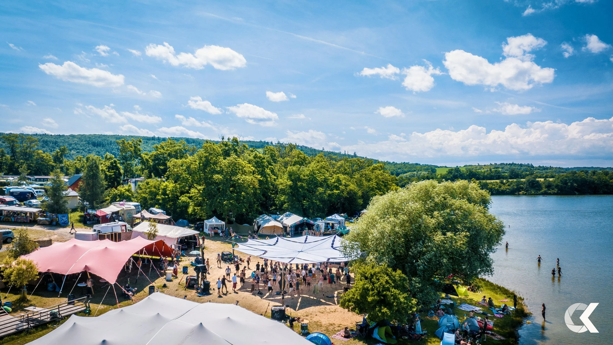 An outdoor lakeside gathering with tents, people, and trees on a sunny day.