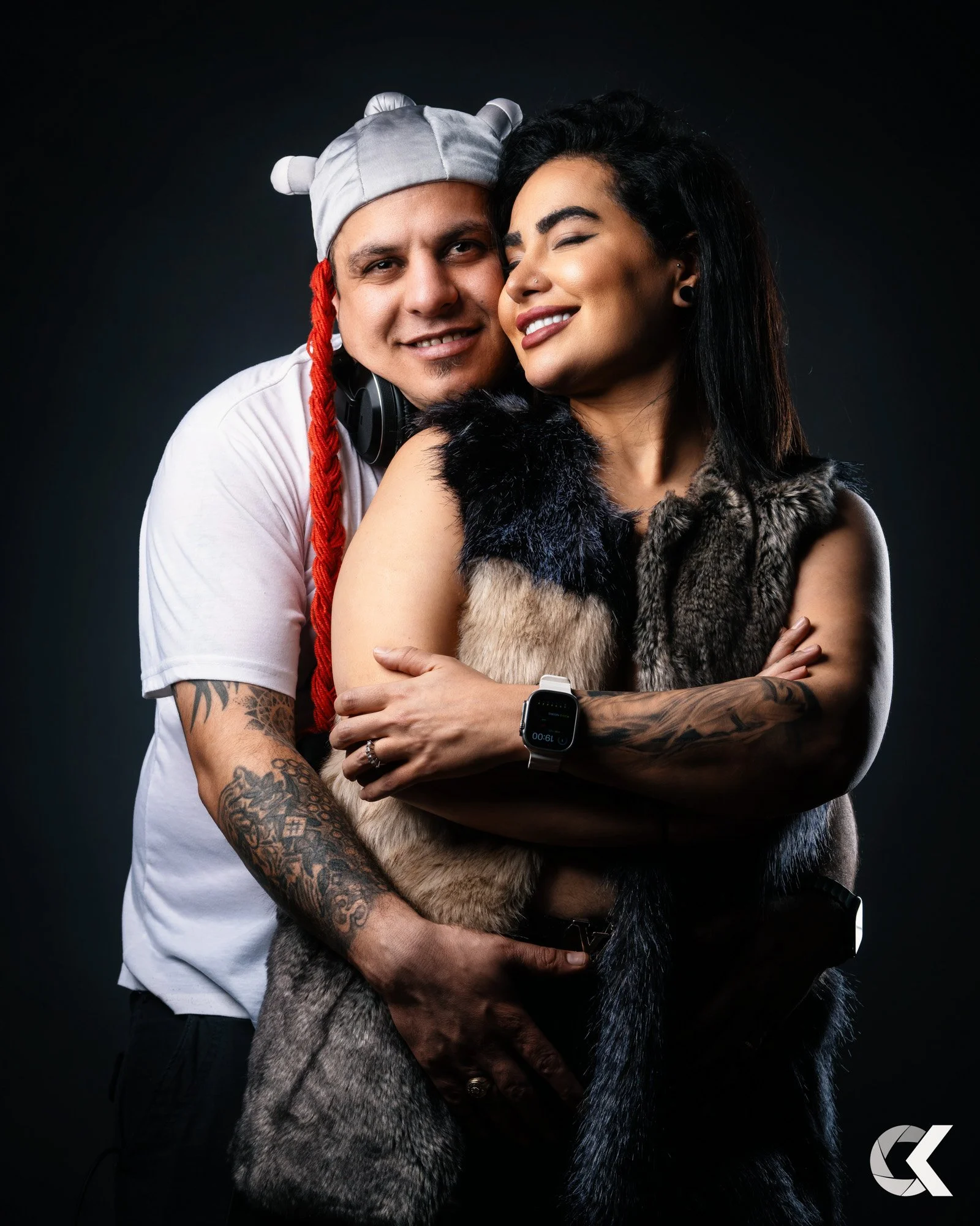 A man and woman hugging, both wearing smartwatches, against a dark background. The man is wearing a gray hat with ears and red braided hair, and has tattoos on his arm. The woman has black hair, tattoos on her arm, and is wearing a fur vest.