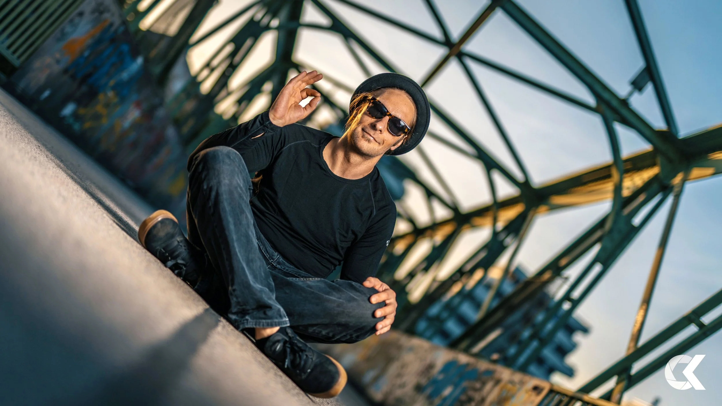 A man sitting on the ground beneath a large metal structure, wearing sunglasses, a black hat, black shirt, and jeans, making an OK gesture with his hand during sunset.